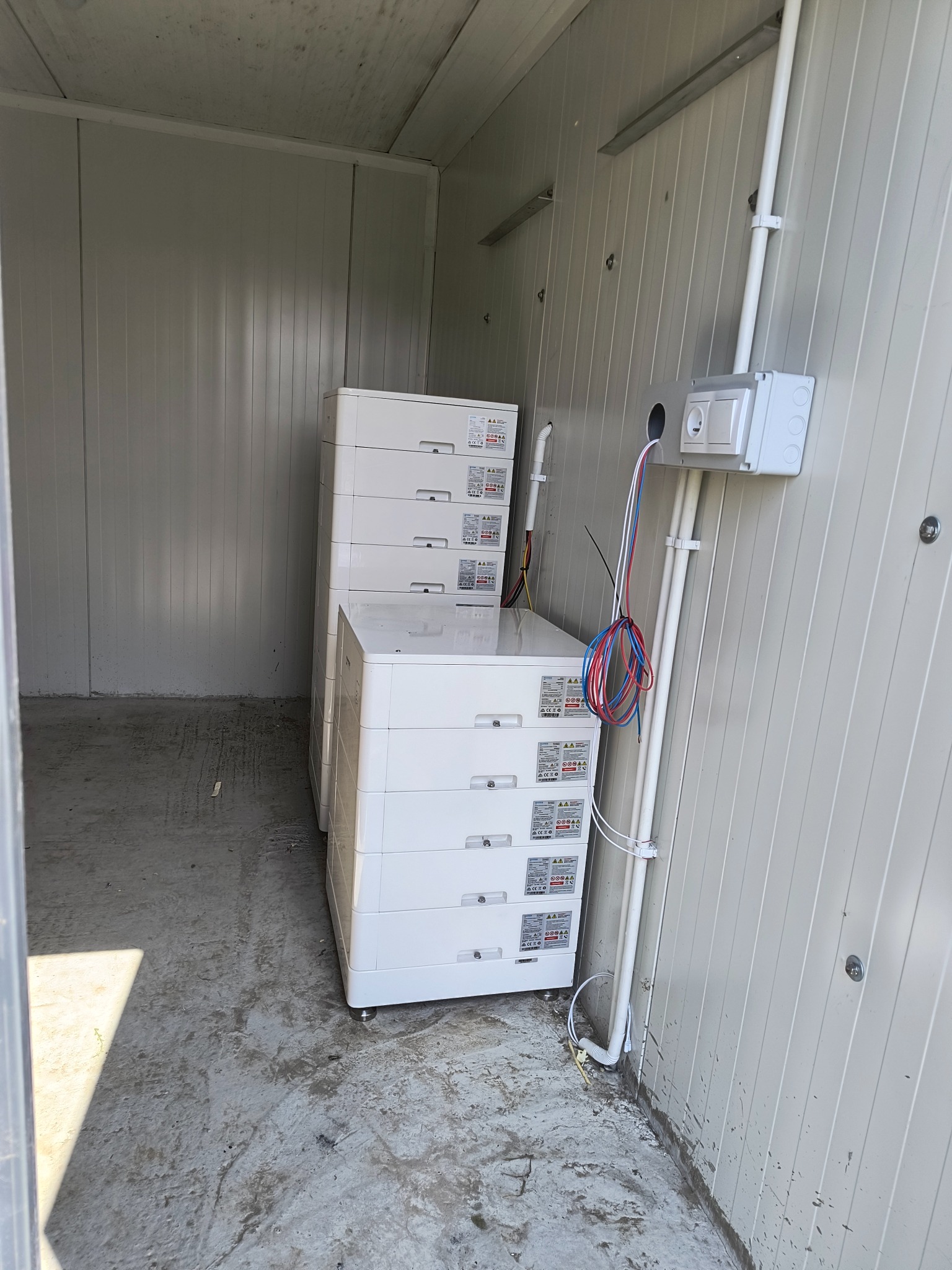 Białe moduły bateryjne w pomieszczeniu technicznym z instalacją elektryczną. Widoczne przewody i gniazdko. Szare ściany i podłoga. Baterie fotowoltaiczne.