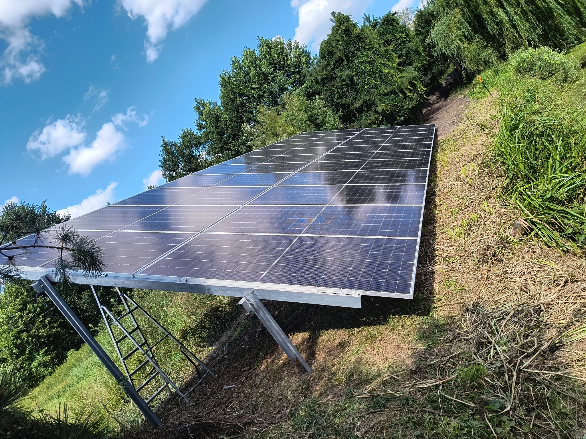 Panel fotowoltaiczny na stelażu, zamontowany na zboczu, z drabiną opartą o konstrukcję. Błękitne niebo z chmurami i zieleń drzew w tle. Ujęcie z dołu.