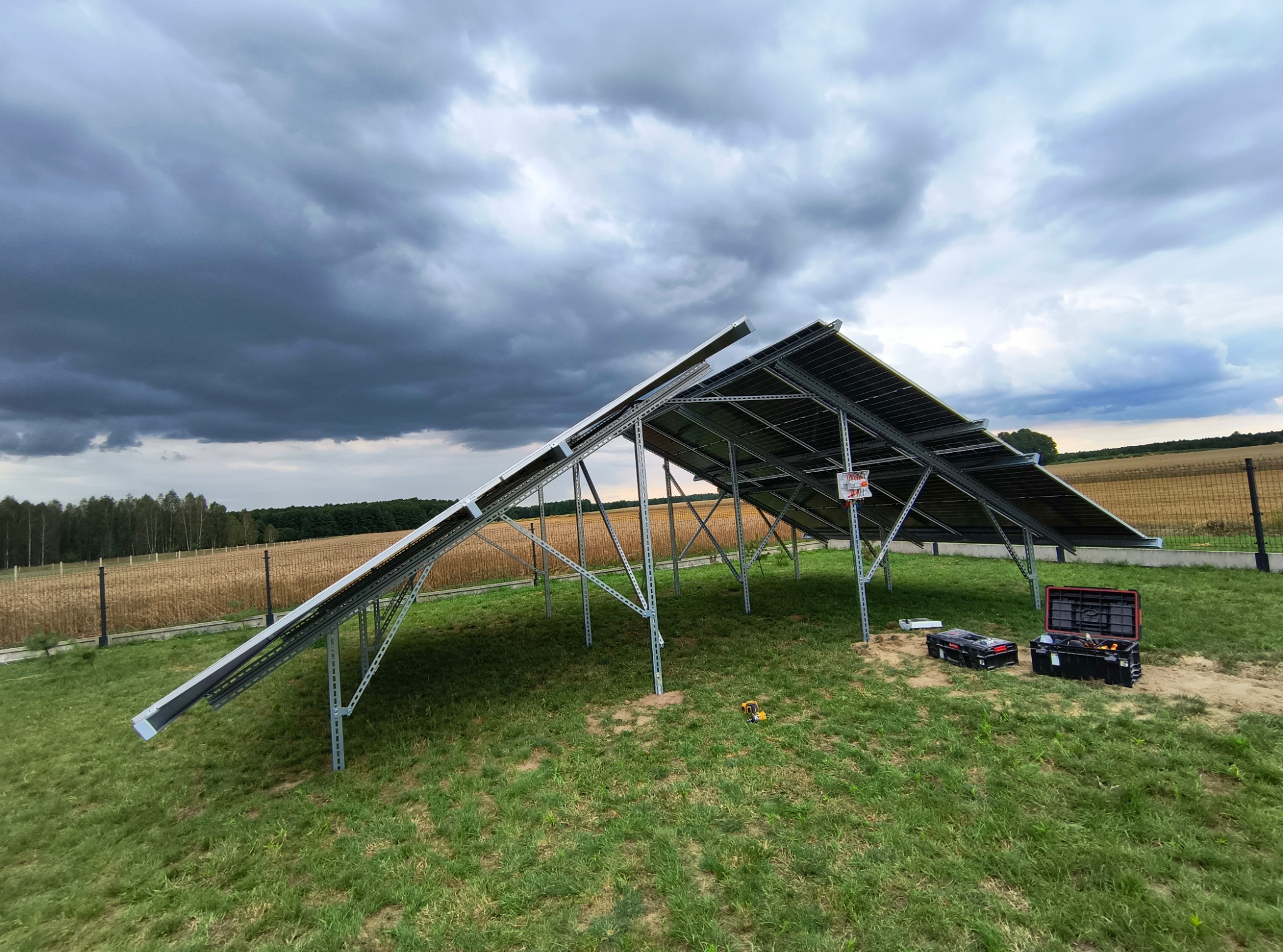 Montaż konstrukcji pod panele fotowoltaiczne na trawie, widoczne narzędzia i pochmurne niebo. Częściowo zamontowane panele. Widok na pole i las w tle.