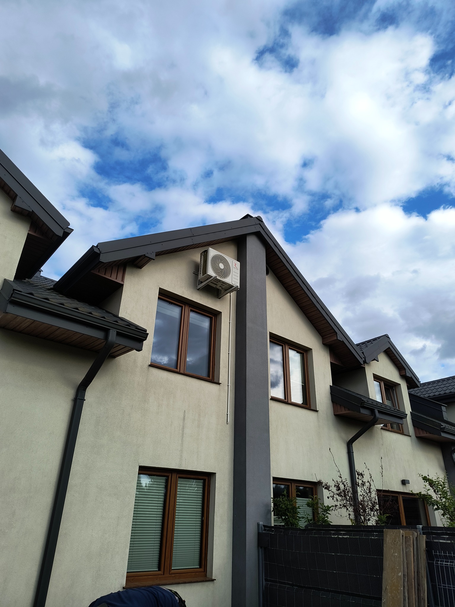 Biała jednostka klimatyzacyjna zamontowana na elewacji budynku mieszkalnego z brązowymi oknami i ciemną dachówką na tle błękitnego nieba z chmurami.