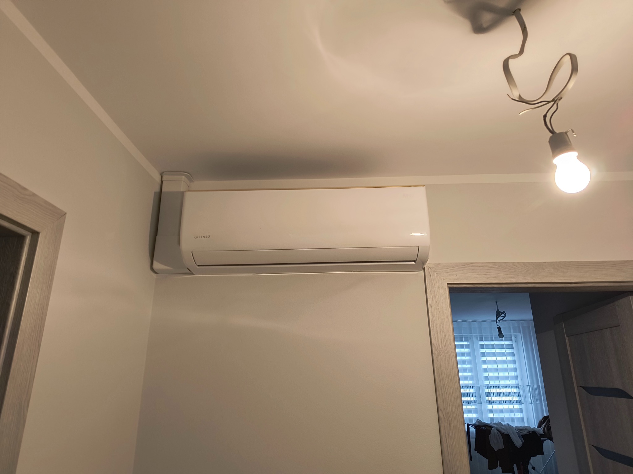 Biała klimatyzacja ścienna marki Itenso, zamontowana w jasnym pomieszczeniu. Widoczny fragment sufitu z lampą bez klosza i fragment drzwi z ościeżnicą.