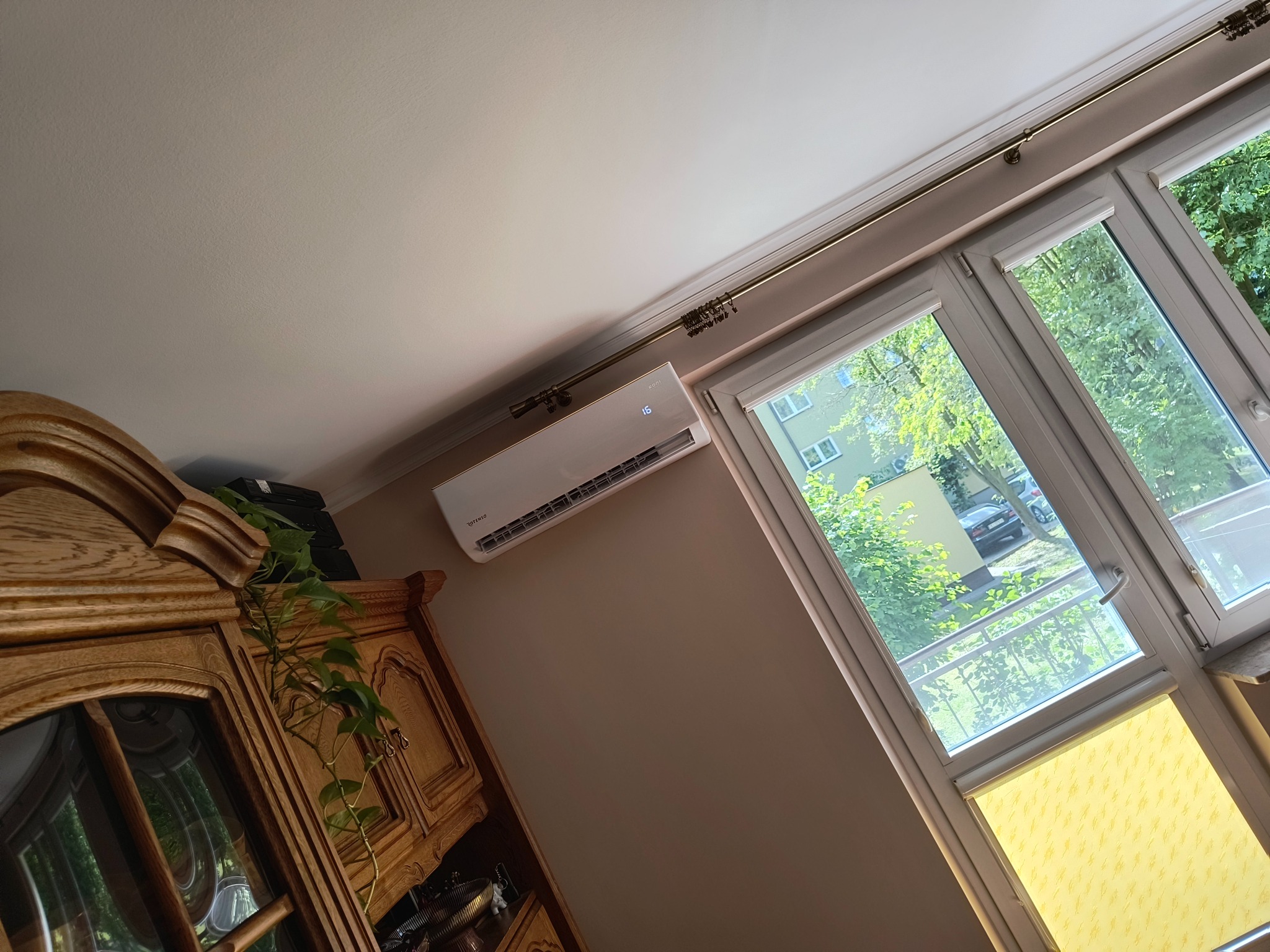 Nowoczesny klimatyzator ścienny, zamontowany w jasnym pokoju z oknem i drewnianą komodą. Widoczny wyświetlacz temperatury. Ujęcie z dołu, podkreślające instalację.