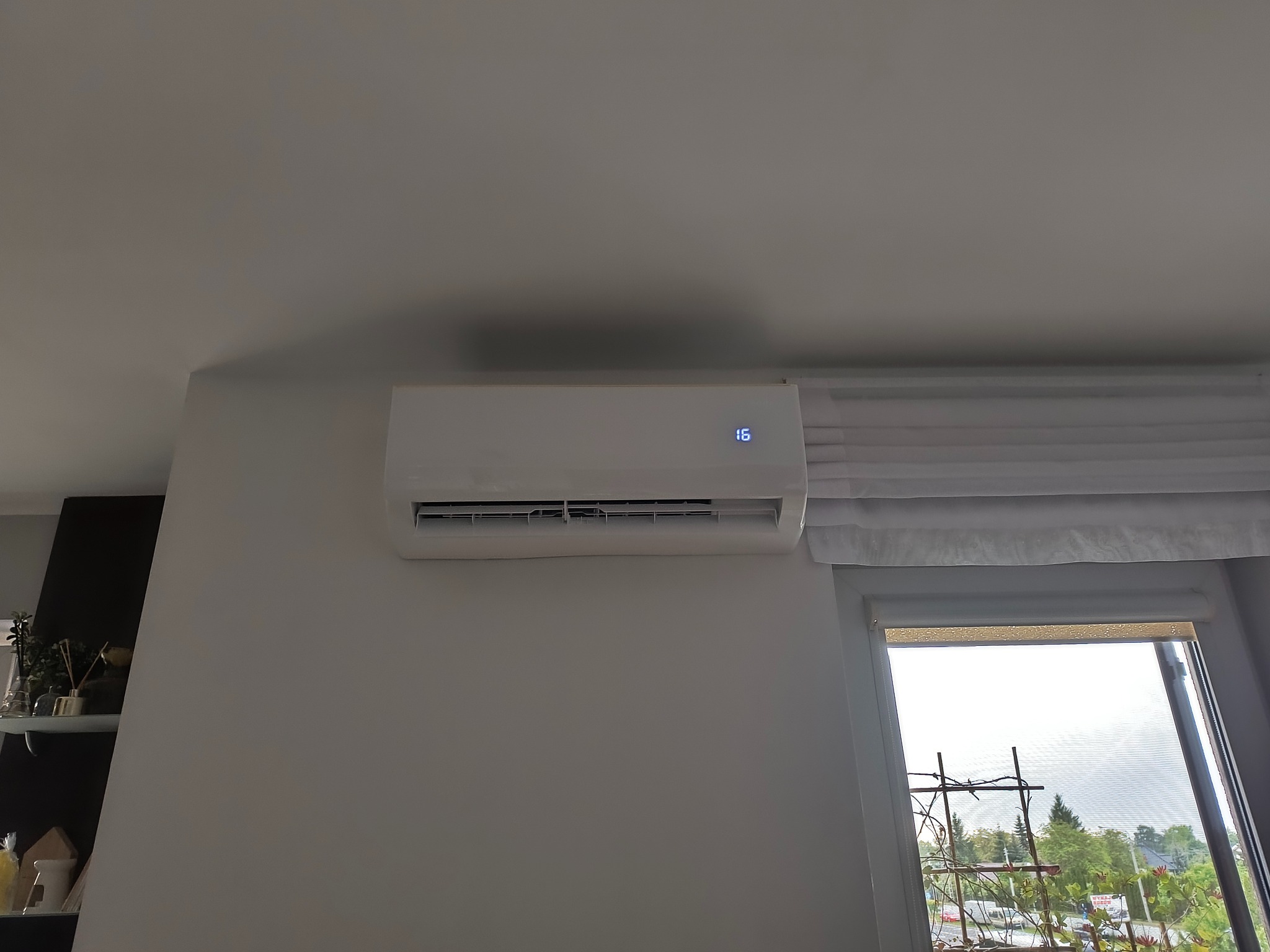 Biały klimatyzator ścienny z wyświetlaczem temperatury (16°C) zamontowany w jasnym wnętrzu, obok okna z widokiem na zieleń. Minimalistyczny design.