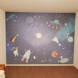Guru Tapetowania - Pokój dziecięcy po tapetowaniu: ściana z motywem kosmosu (planety, gwiazdy, rakiety, astronauta) na ciemnoniebieskim tle. Widoczna część podłogi i mebla.