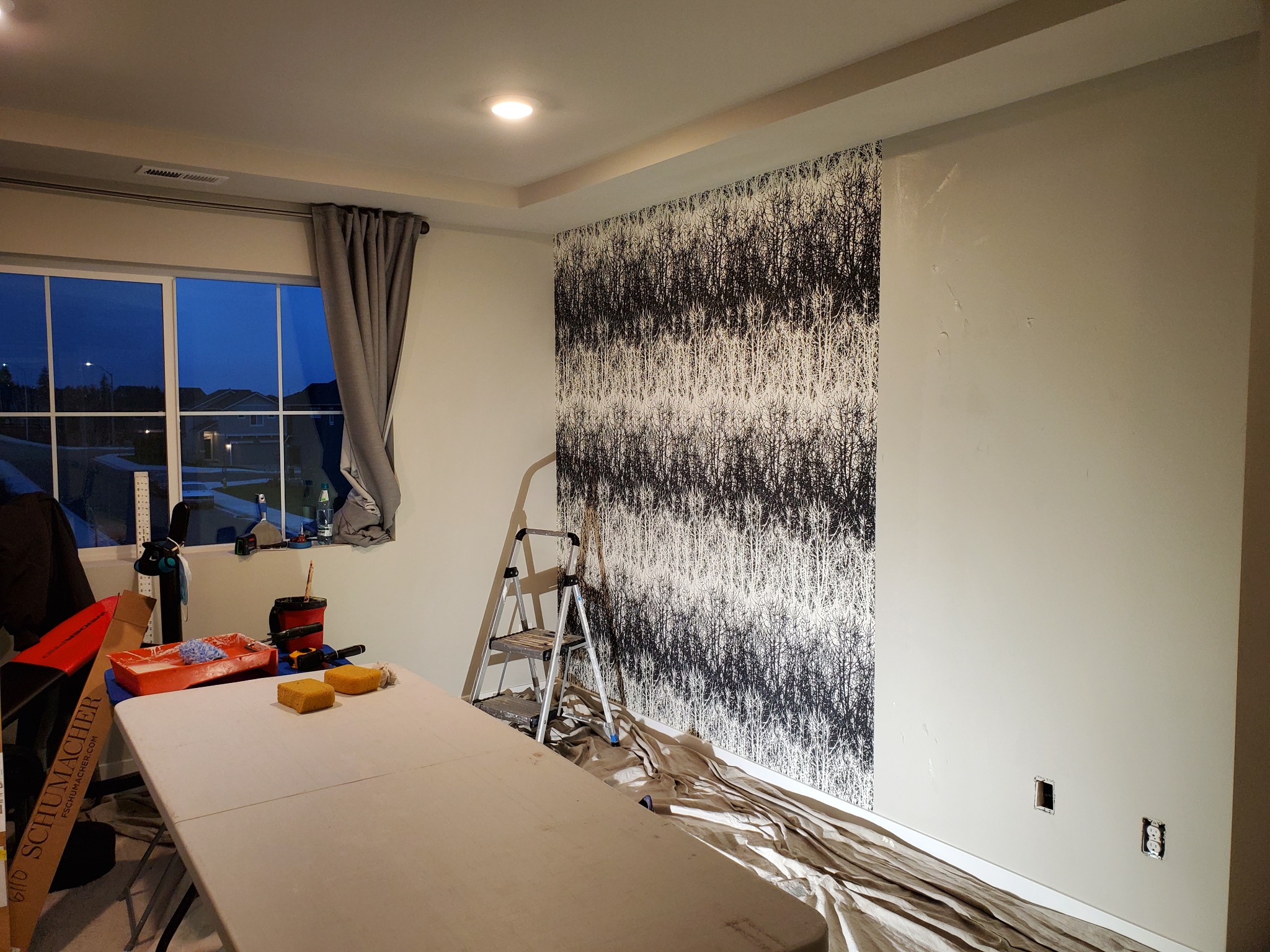 Tapetowanie ściany w pokoju z oknem. Widoczna drabina, stół z narzędziami i wzorzysta tapeta w czarno-białe drzewa. Przy oknie szare zasłony.