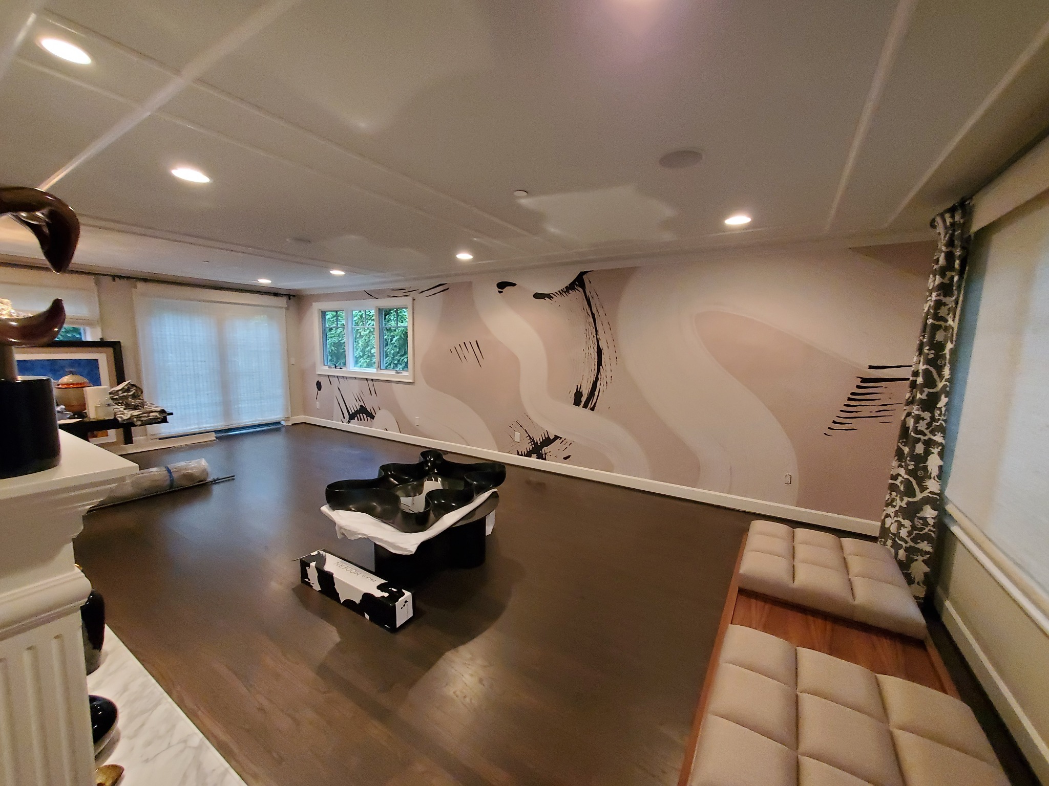 Przestronny salon z ciemną podłogą i oryginalną, abstrakcyjną dekoracją ścienną w odcieniach beżu i czerni. Nowoczesne meble i jasne światło podkreślają unikalny charakter wnętrza.