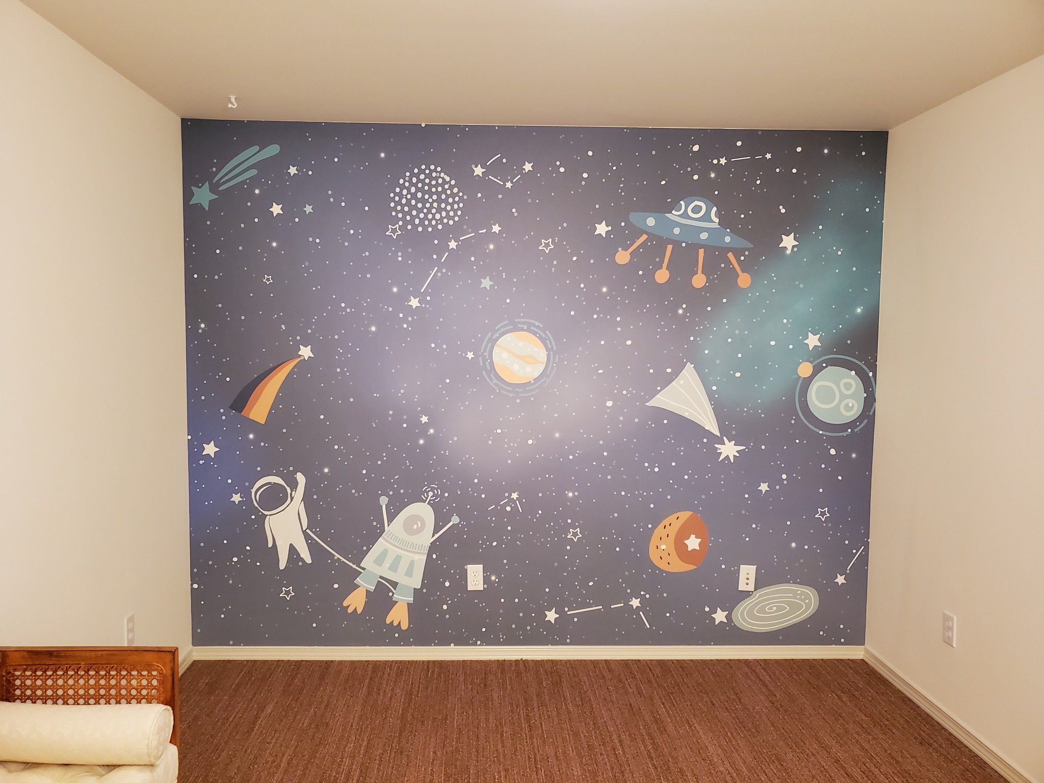 Pokój dziecięcy po tapetowaniu: ściana z motywem kosmosu (planety, gwiazdy, rakiety, astronauta) na ciemnoniebieskim tle. Widoczna część podłogi i mebla.