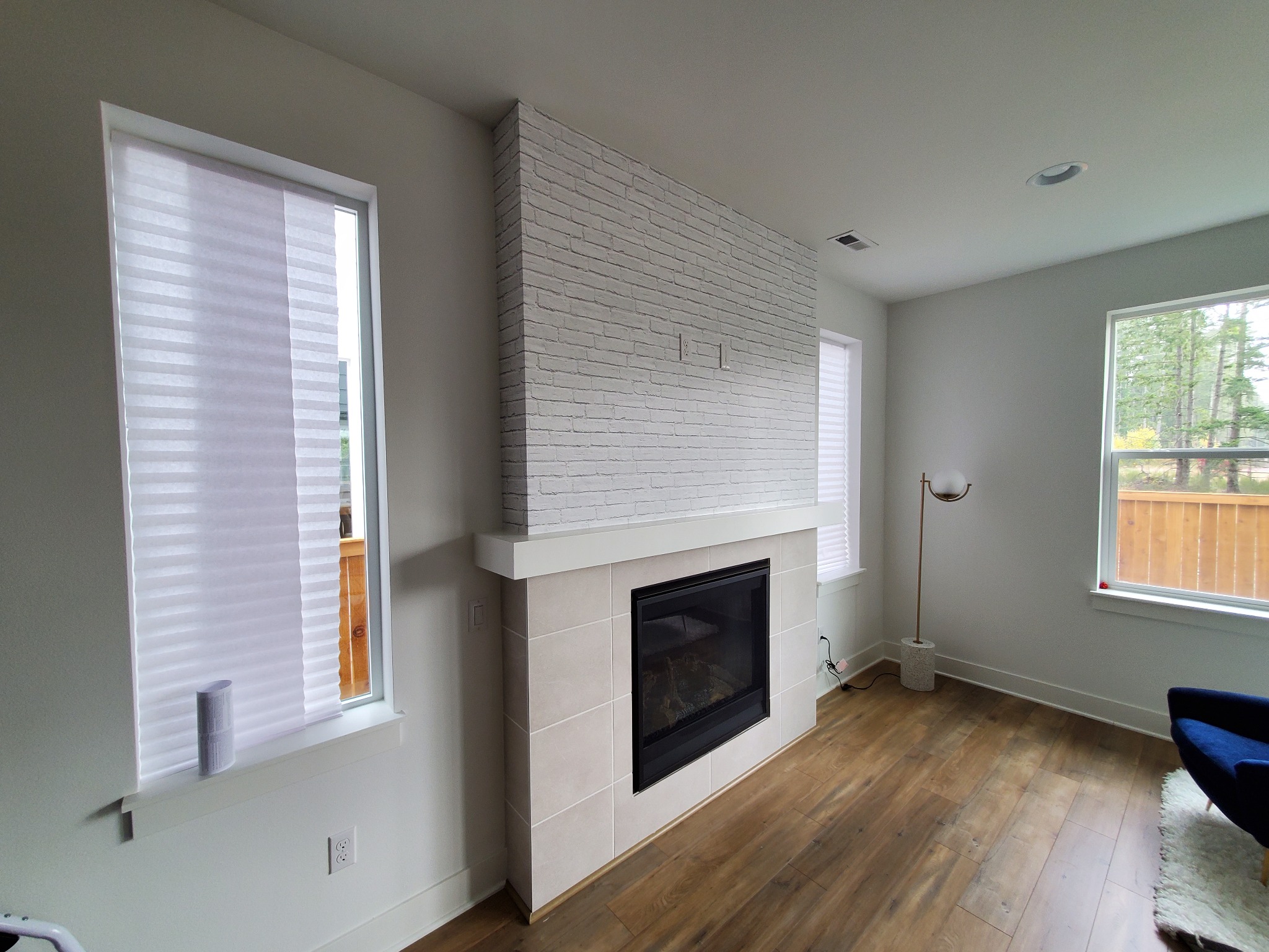 Salon z kominkiem obłożonym jasną cegłą i płytkami, drewniana podłoga, okno z żaluzjami plisowanymi. Minimalistyczny design, nowoczesne wykończenie.