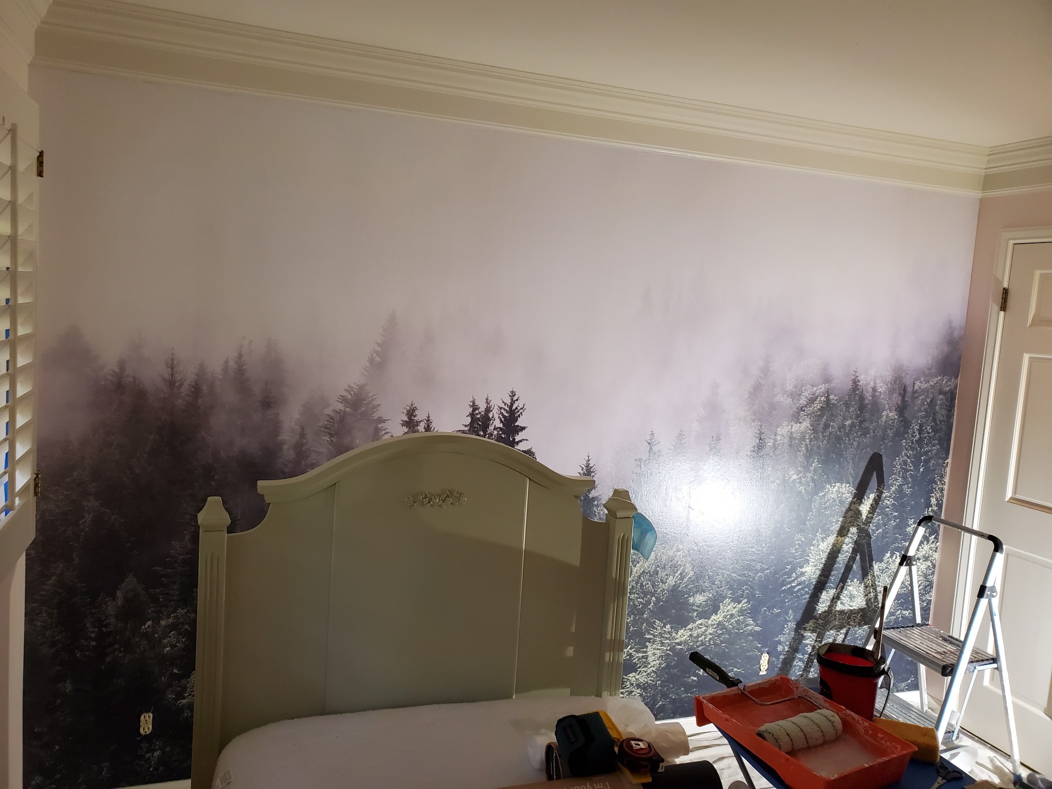 Pokój po tapetowaniu fototapetą z motywem mglistego lasu. Widoczna drabina, wałek i kuweta malarska. Wezgłowie łóżka zasłania część ściany.
