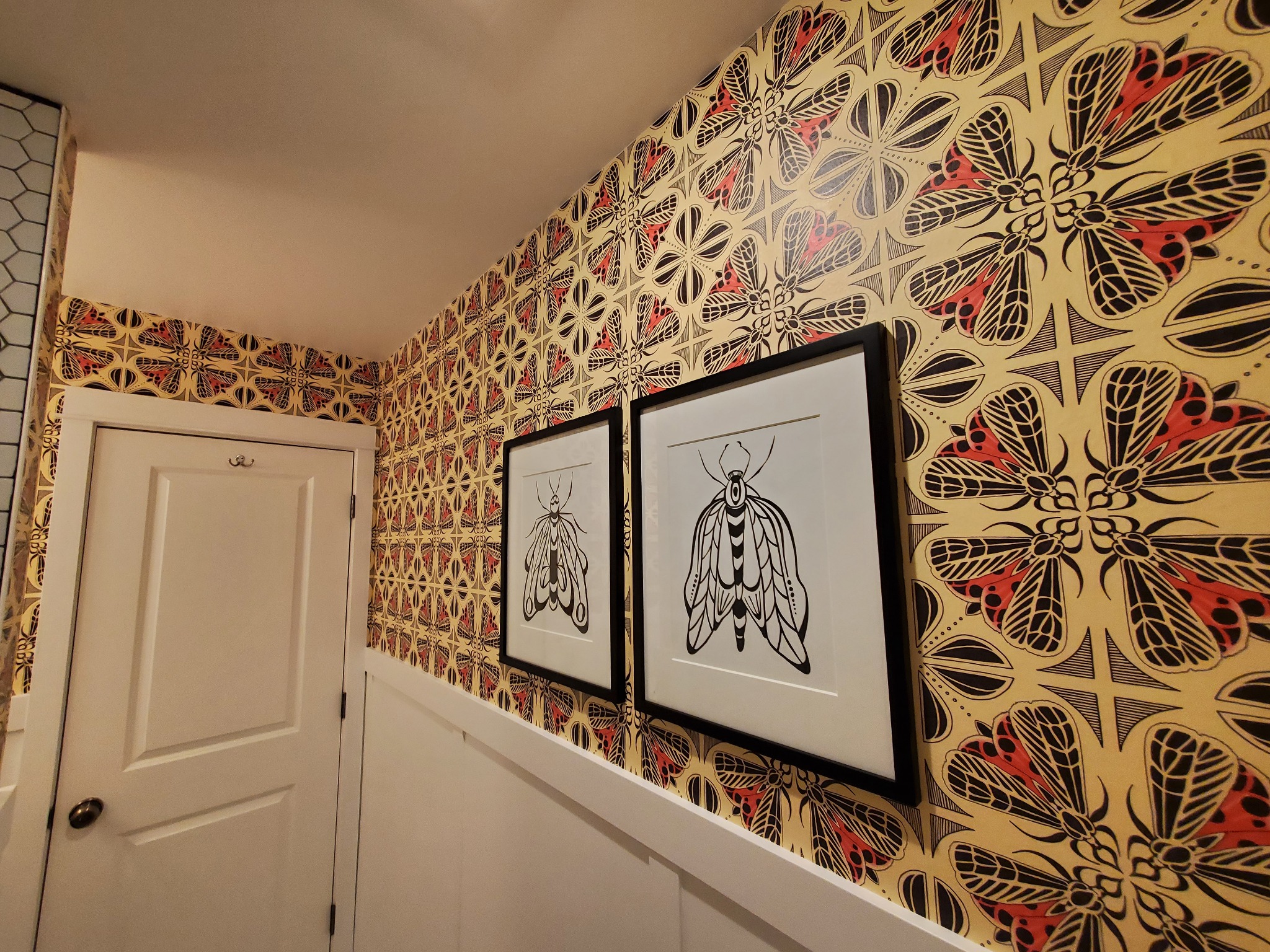 Wzorzysta tapeta w motyle w korytarzu, z dwoma obrazami owadów w czarnych ramkach. Białe drzwi po lewej stronie. Ujęcie z dołu, podkreślające wzór i detale.
