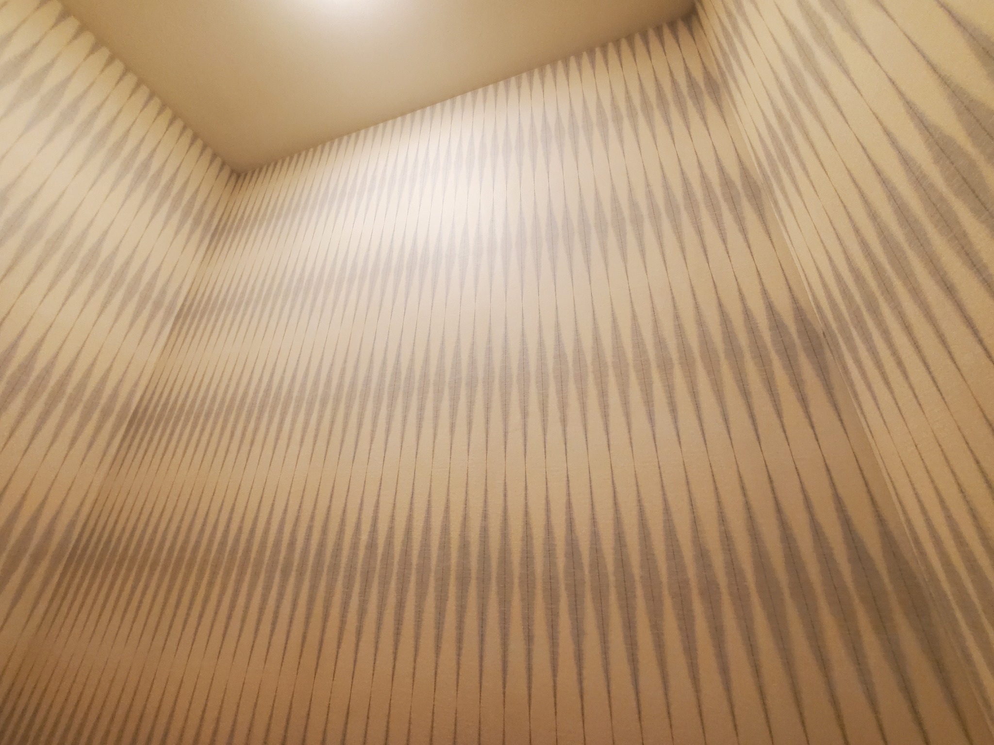 Wnętrze z tapetą w geometryczny wzór w odcieniach beżu i szarości. Widok na ścianę i fragment sufitu, oświetlonego punktowym światłem.