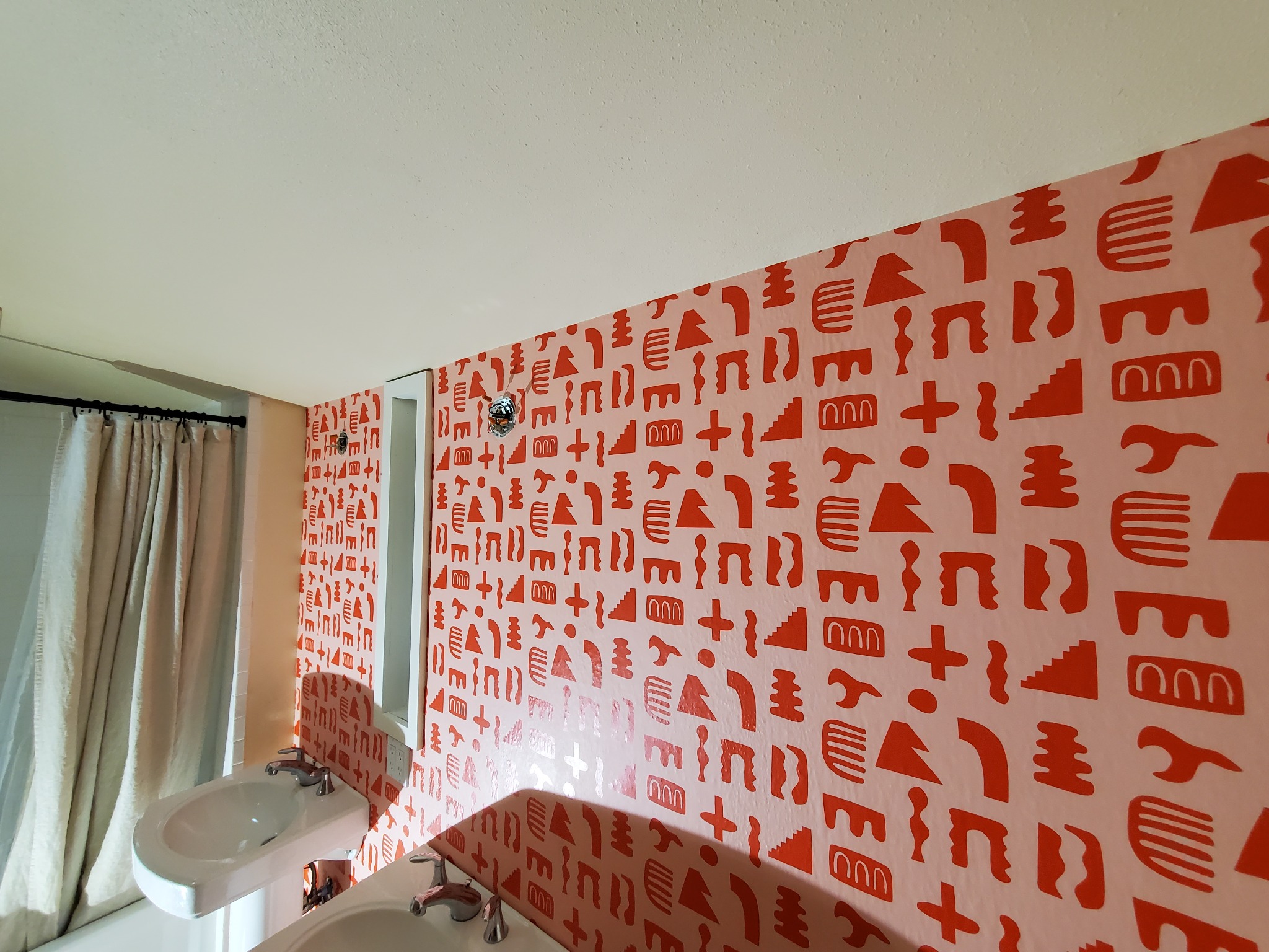 Łazienka po tapetowaniu z wzorem geometrycznym w odcieniach różu i czerwieni. Dwie umywalki i zasłona prysznicowa w tle. Nietypowy deseń tapety.