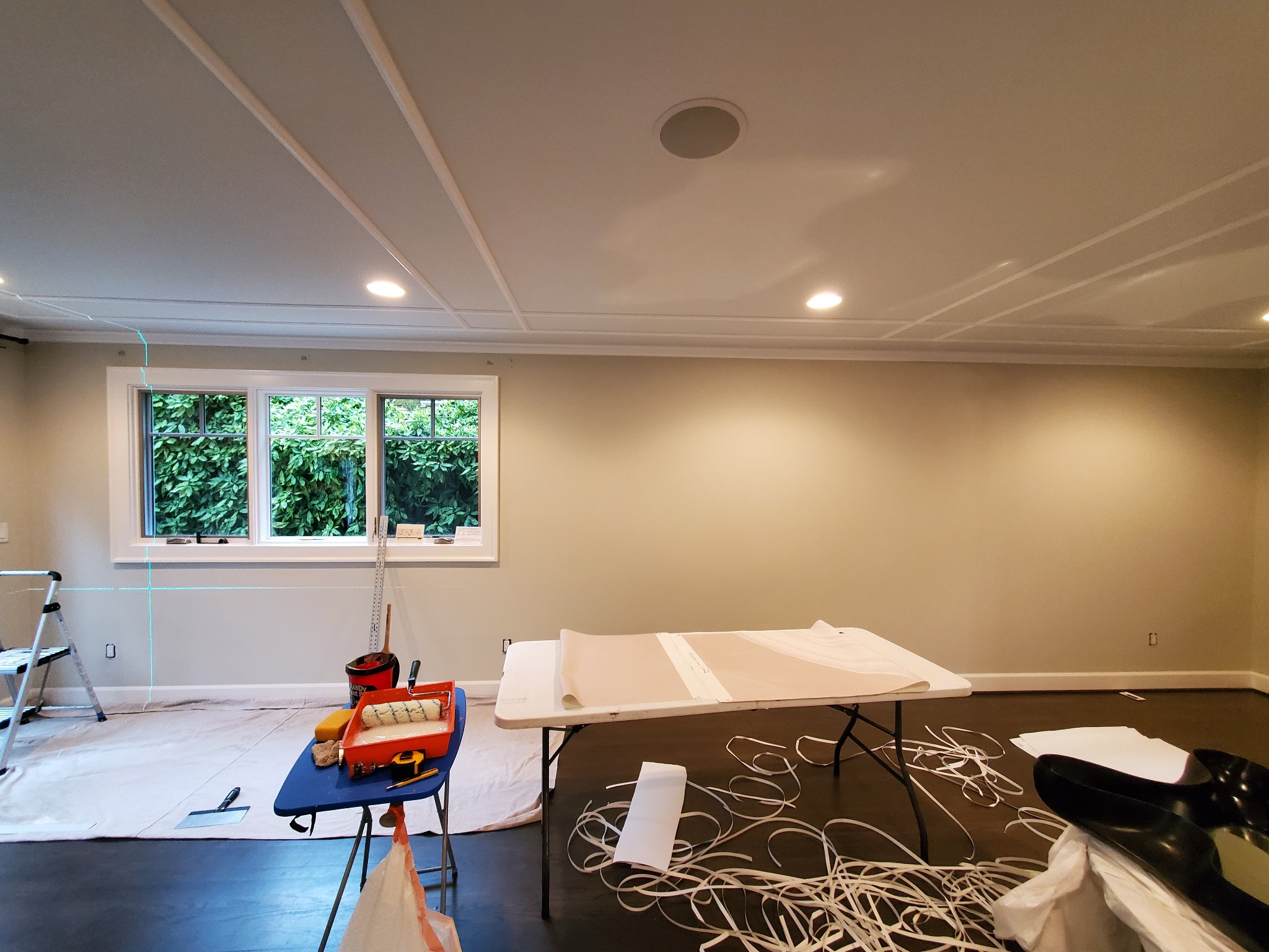 Pokój w trakcie przygotowania do tapetowania: stół z tapetą, narzędzia malarskie, drabina, zabezpieczona podłoga. Widoczne listwy dekoracyjne na suficie.