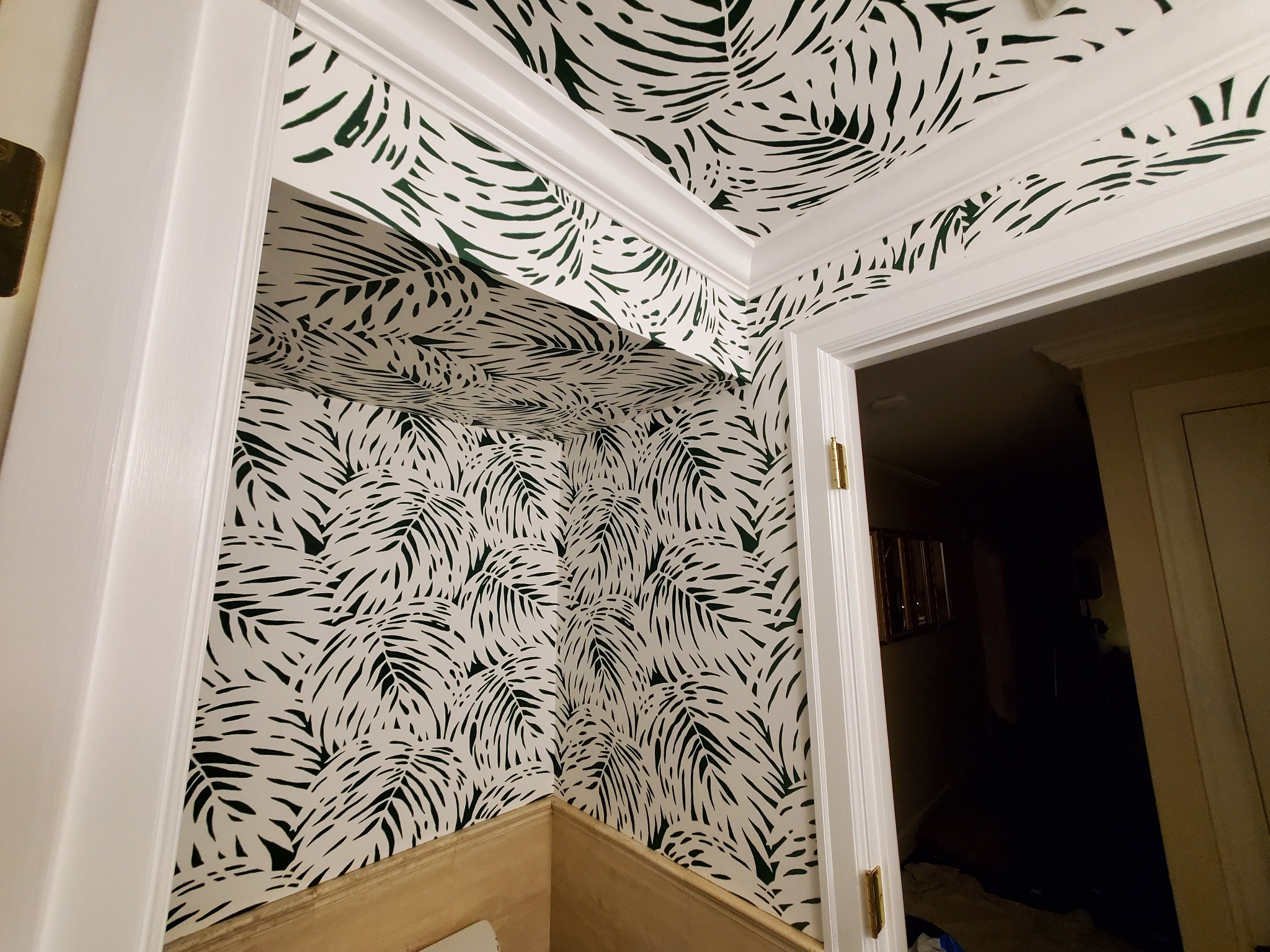 Wnętrze po tapetowaniu: ściany i sufit pokryte tapetą w zielone liście na białym tle, widoczne białe listwy przypodłogowe i framuga drzwi. Dekoracyjny wzór liści palmowych.