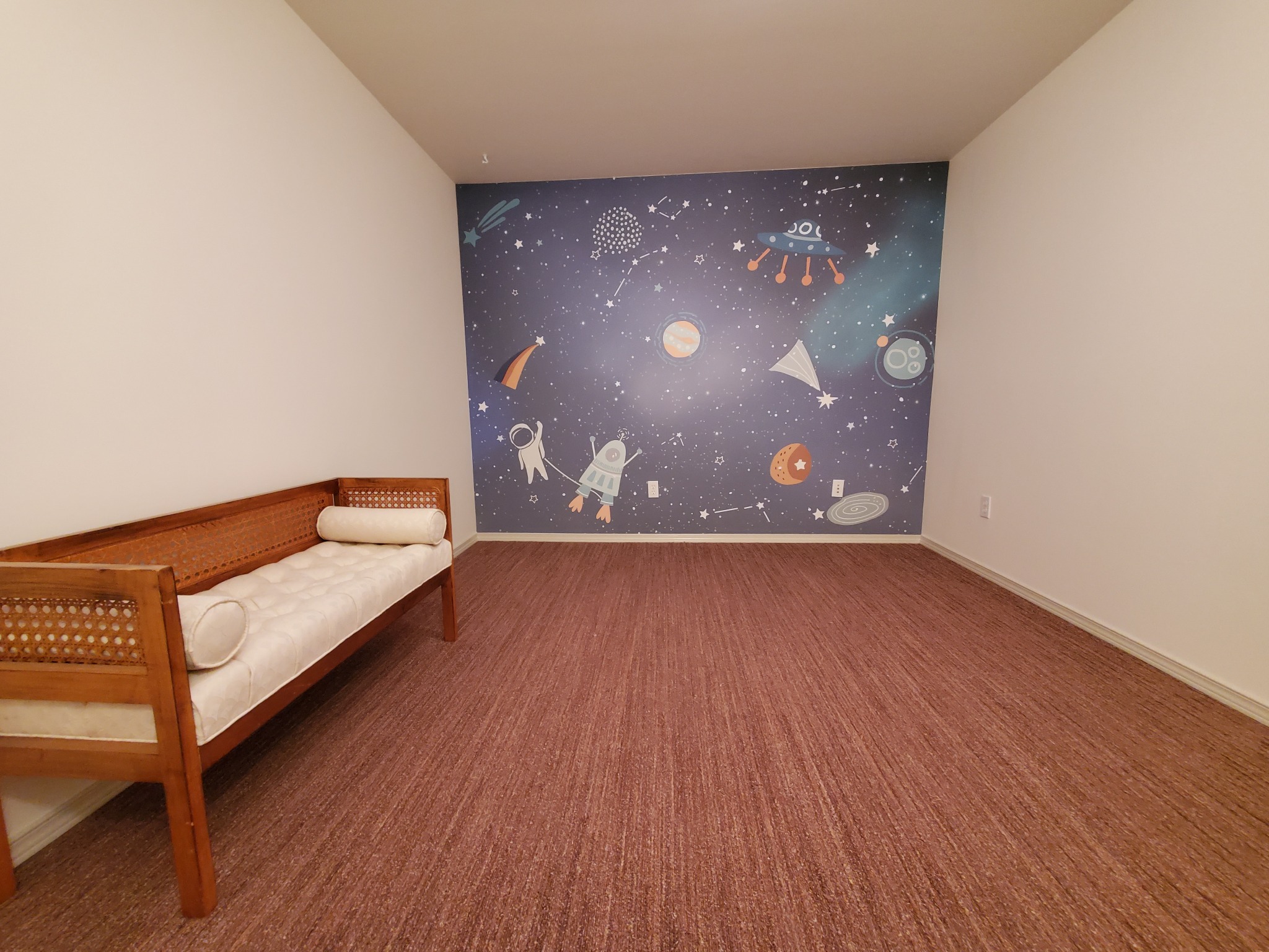 Pokój po tapetowaniu z motywem kosmosu na ścianie. Wnętrze dopełnia drewniana kanapa z jasną tapicerką i brązowa wykładzina dywanowa.