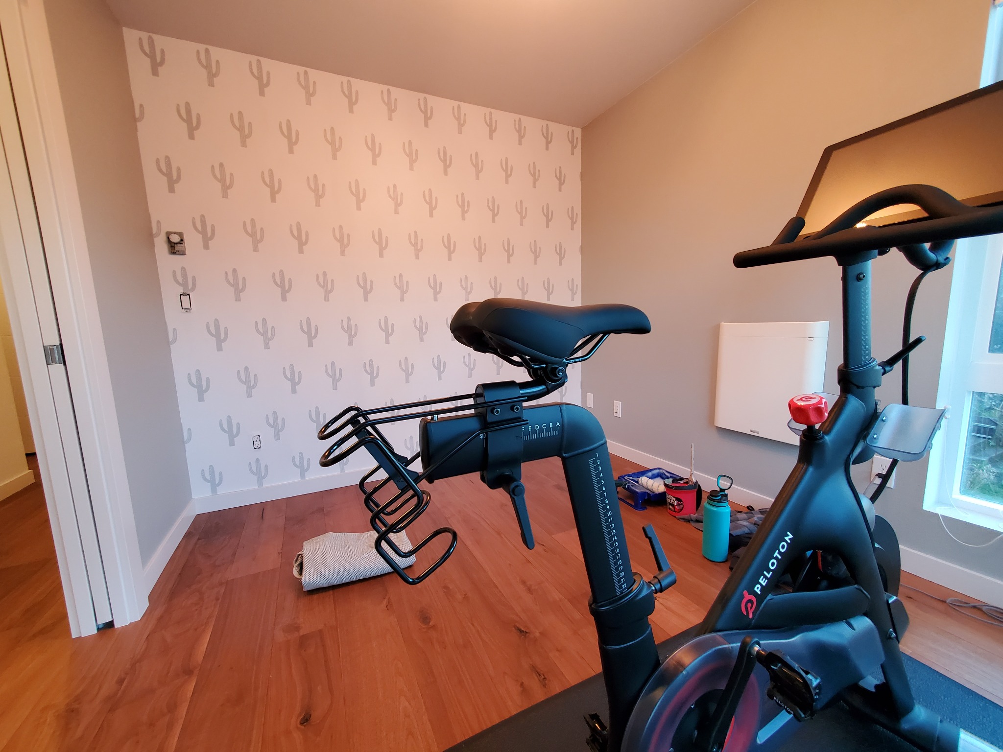 Pokój z drewnianą podłogą i ścianą w kaktusy, na której stoi czarny rower treningowy Peloton. Widoczne akcesoria do ćwiczeń i okno z naturalnym światłem.