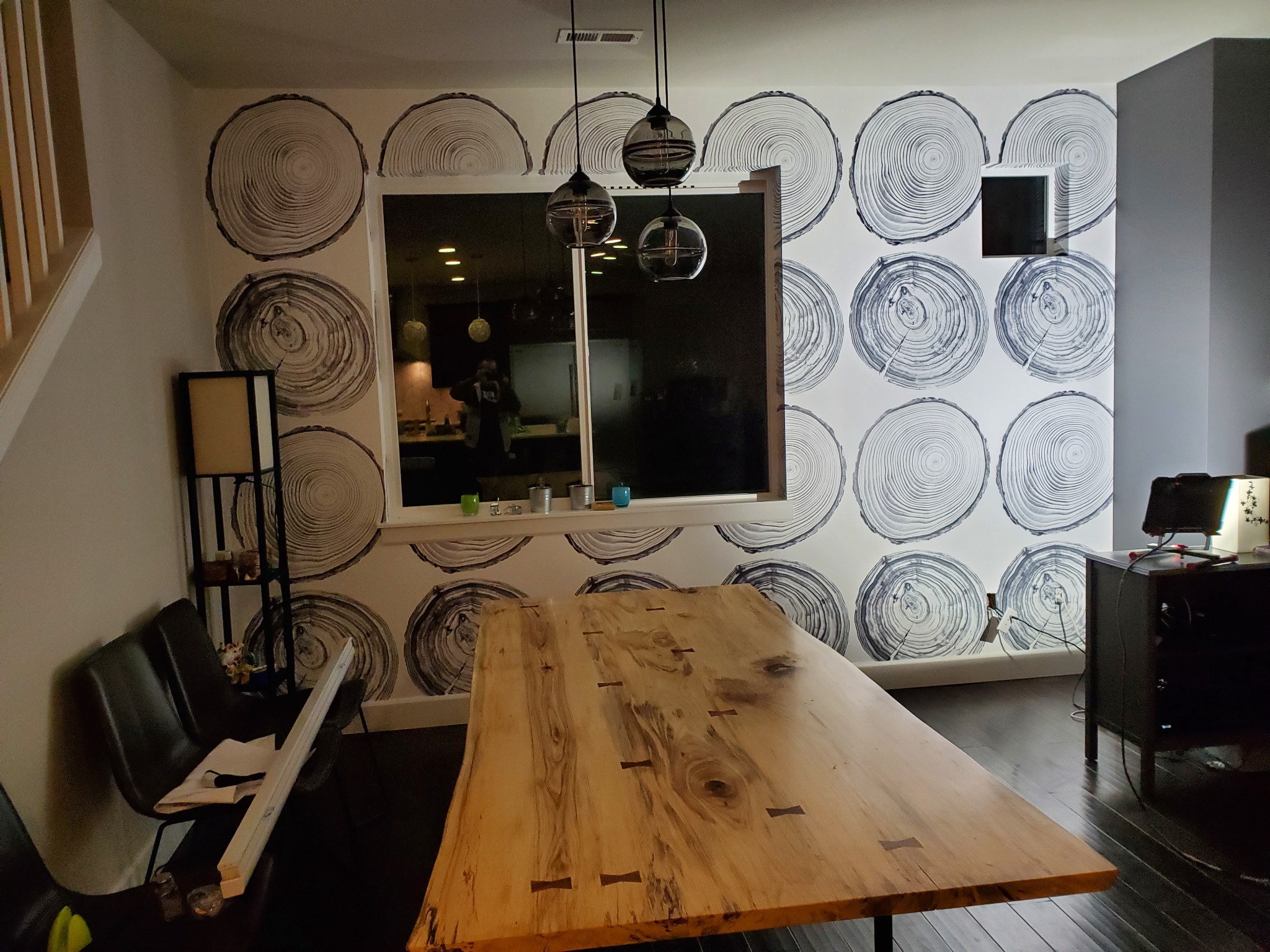 Ściana z tapetą w motywie przekroju pnia drzewa, drewniany stół z widocznymi łączeniami, nowoczesne lampy wiszące i czarne krzesła w tle.