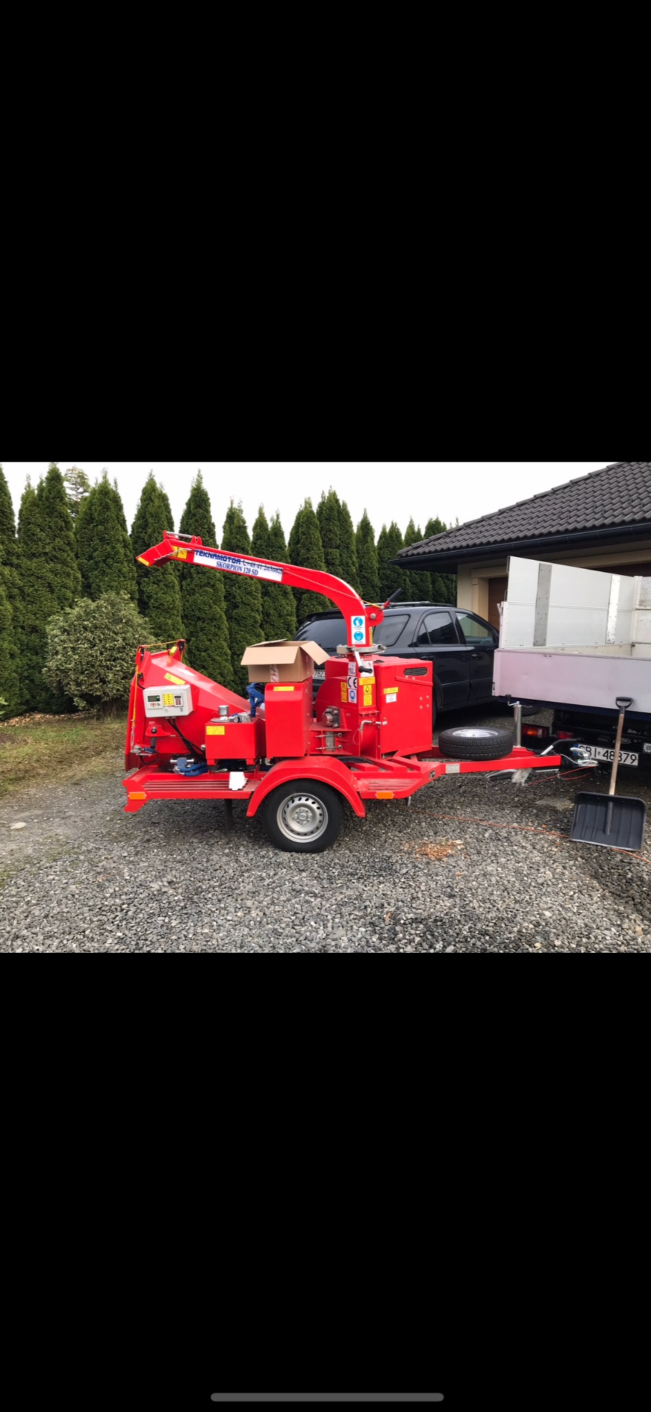 Czerwona, mobilna rębarka do gałęzi Teknamotor Skorpion 120 SD na żwirowym podłożu, z przyczepionym do niej samochodem dostawczym i łopatą. W tle tuje i dom.