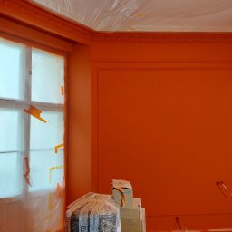 Viacheslav Dunda - Pokój po malowaniu na pomarańczowo, zabezpieczone okno folią, widoczne kartony i narzędzia malarskie. Ściany z dekoracyjnymi listwami.