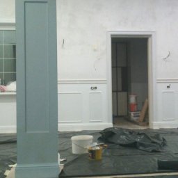 Viacheslav Dunda - Malowanie kolumny w odcieniach szarości w pomieszczeniu z białymi panelami ściennymi. Widoczne narzędzia malarskie i zabezpieczenia podłogi.