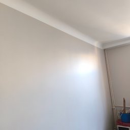 Viacheslav Dunda - Jasna ściana po malowaniu z dekoracyjną listwą przysufitową. W rogu widoczne rury grzewcze i akcesoria malarskie, co sugeruje świeżo zakończone prace wykończeniowe.