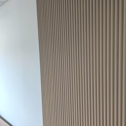 Viacheslav Dunda - Wykończona ściana z dekoracyjnymi, pionowymi panelami w odcieniach beżu, przylegająca do gładkiej, białej ściany. Widoczne detale wykończenia i fragment podłogi.