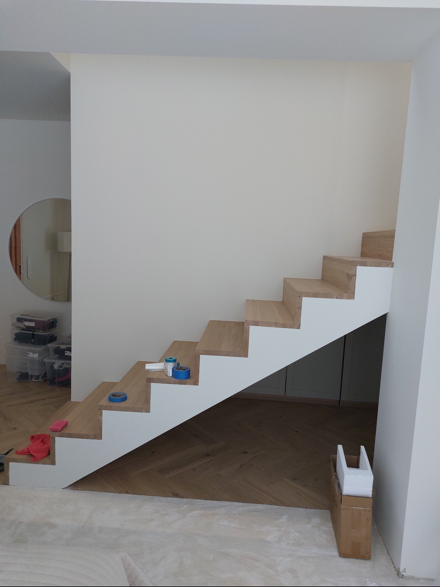 Schody drewniane z białymi podstopnicami w trakcie zabezpieczania taśmą malarską. Widoczne narzędzia i karton. Podłoga zabezpieczona folią malarską.
