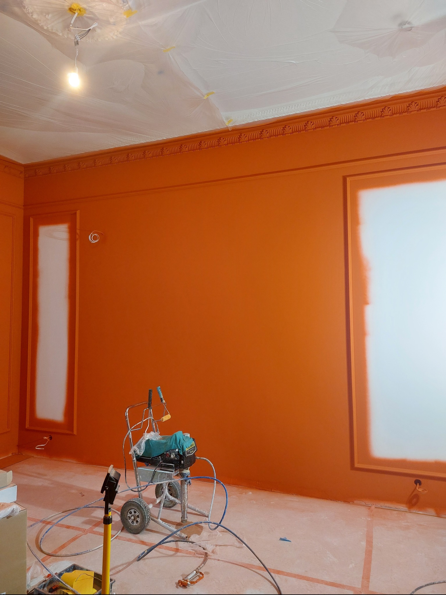 Pokój w trakcie malowania natryskowego na pomarańczowo, z zabezpieczonym sufitem folią i częściowo zamalowanymi wnękami. Widoczny agregat malarski i akcesoria.