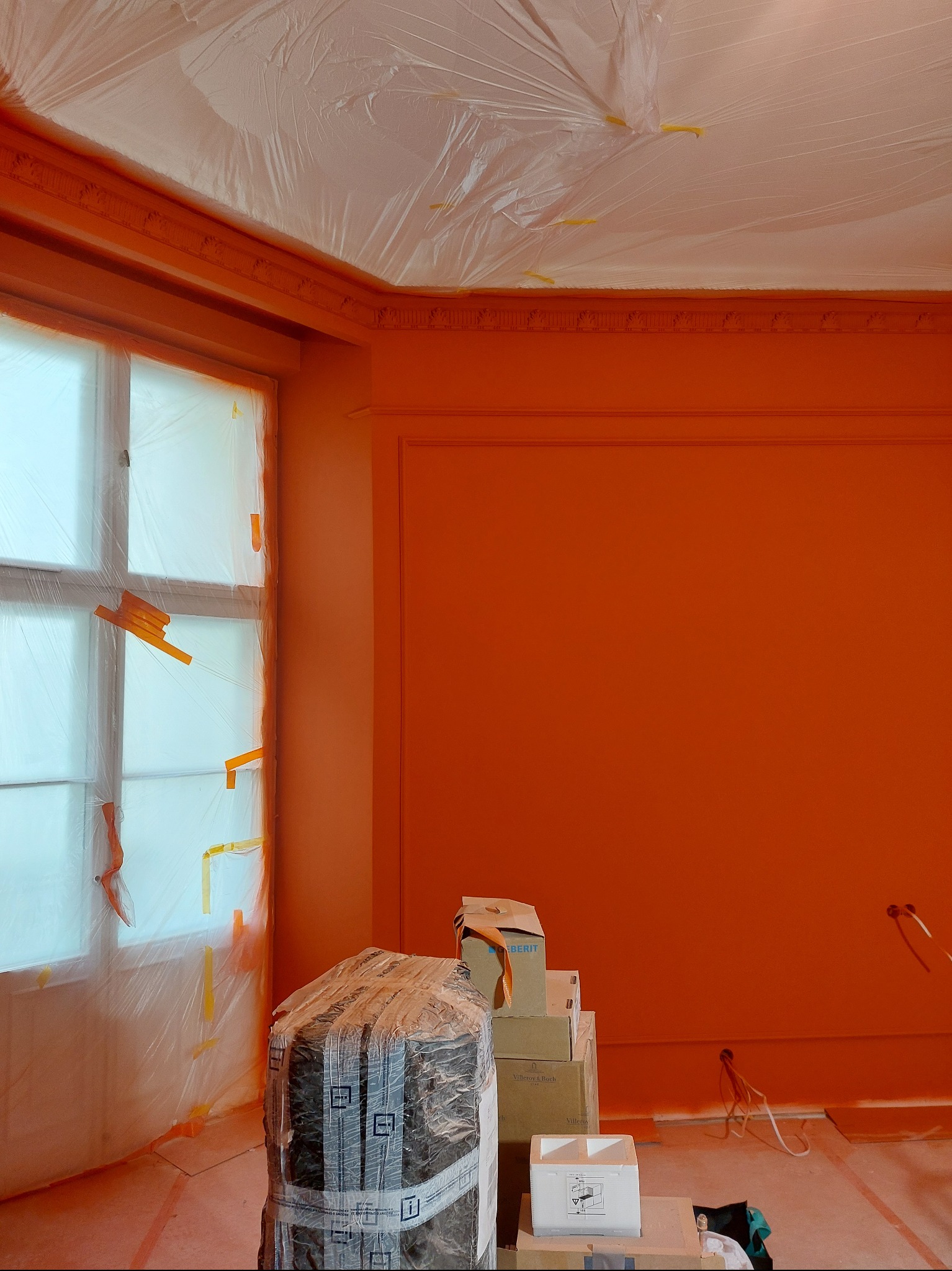 Pokój po malowaniu na pomarańczowo, zabezpieczone okno folią, widoczne kartony i narzędzia malarskie. Ściany z dekoracyjnymi listwami.