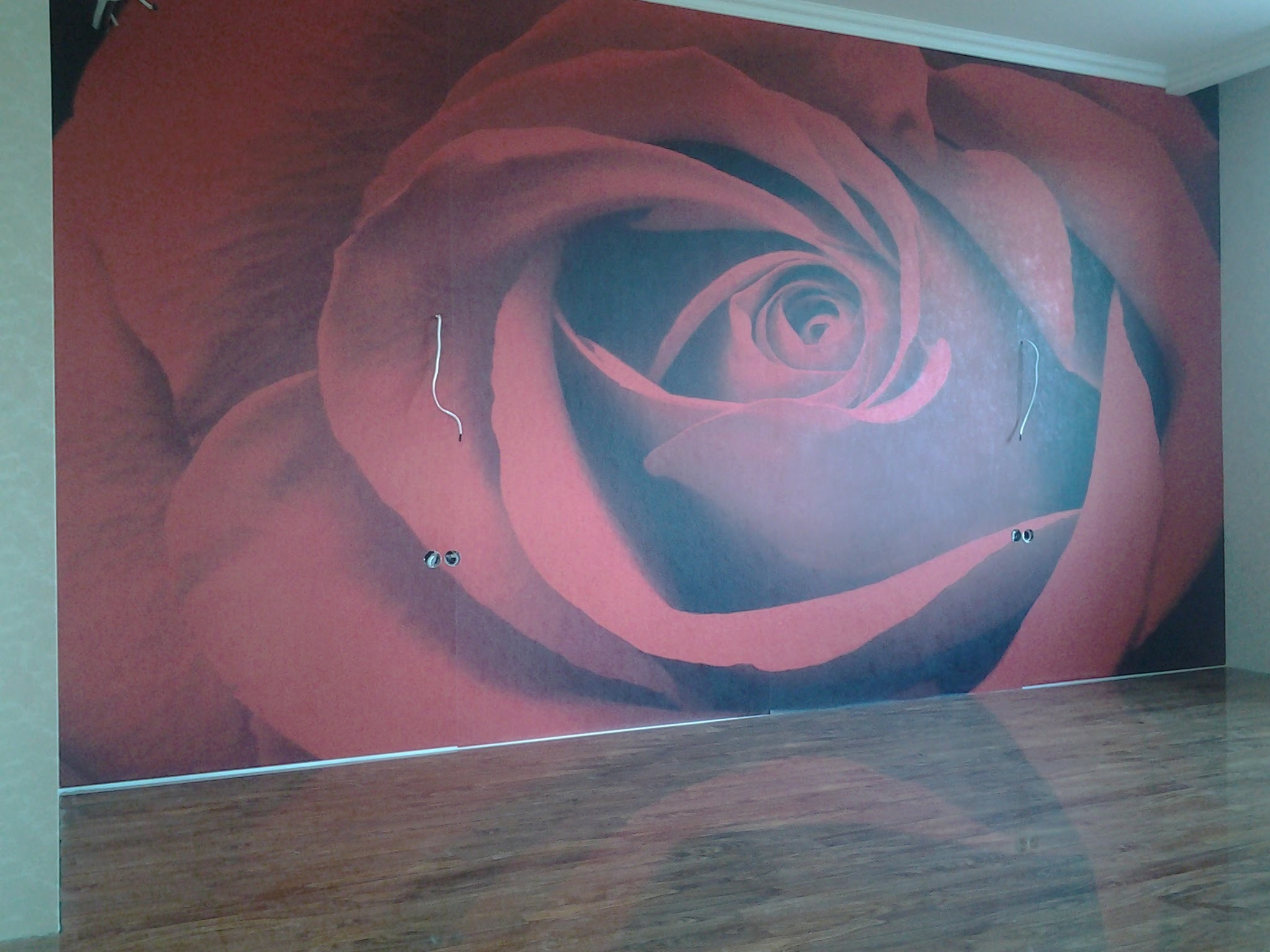 Ściana z fototapetą w motywie czerwonej róży, widoczne gniazdka elektryczne i fragment drewnianej podłogi. Dekoracja wnętrza po remoncie.