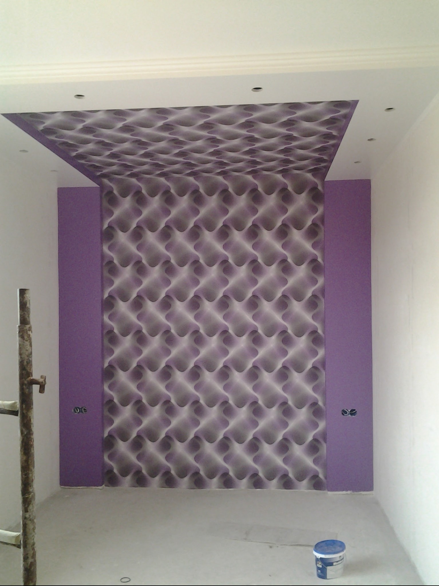 Wnętrze po remoncie: ściana z wzorzystą tapetą w odcieniach fioletu i szarości, obok pionowy pas pomalowany na fioletowo. Widoczny fragment drabiny i wiadro z farbą.
