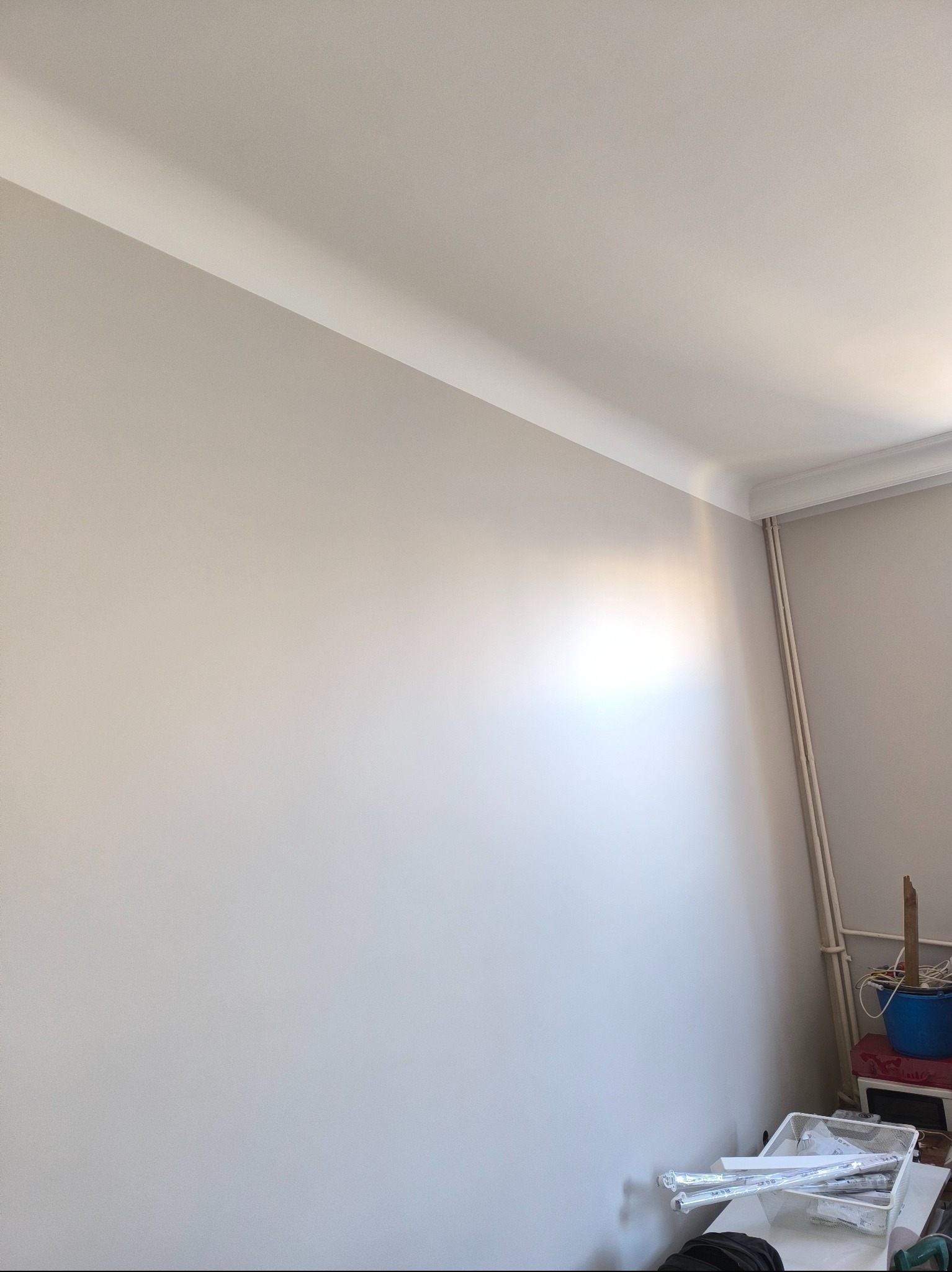 Jasna ściana po malowaniu z dekoracyjną listwą przysufitową. W rogu widoczne rury grzewcze i akcesoria malarskie, co sugeruje świeżo zakończone prace wykończeniowe.