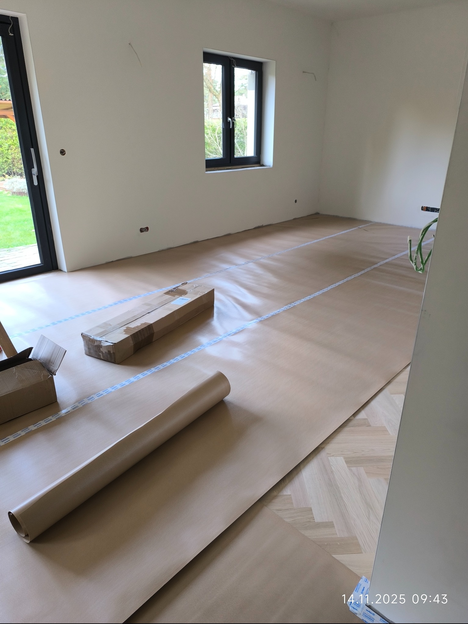 Pokój zabezpieczony brązowym papierem malarskim i taśmą. Widoczne kartony i rolka papieru. Parkiet w jodełkę częściowo zakryty. Przygotowanie do malowania ścian.