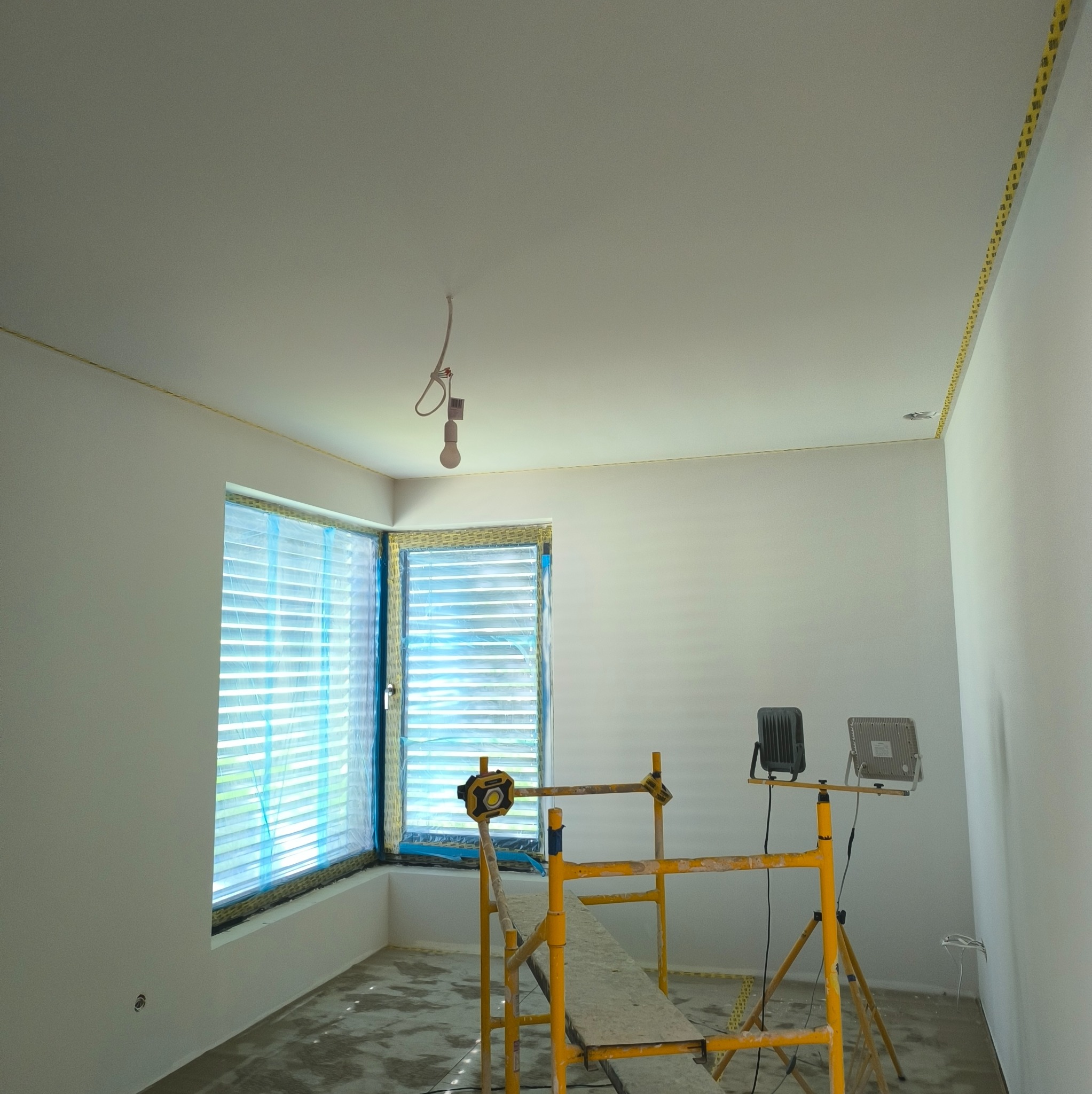 Pokój w trakcie prac wykończeniowych: zabezpieczone okno, ściany przygotowane do malowania, żółte rusztowanie i lampy halogenowe. Widoczny surowy beton na podłodze.