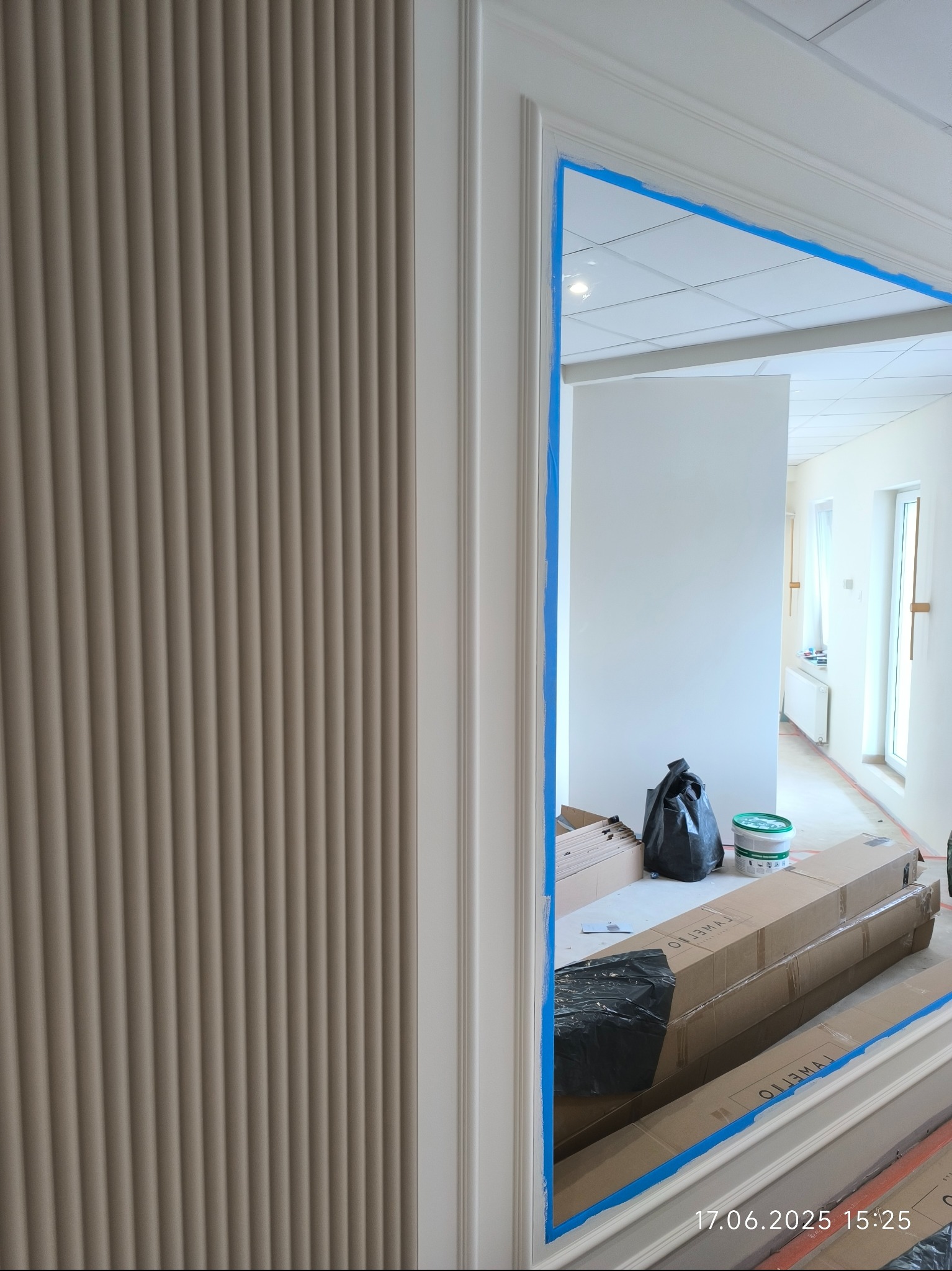 Beżowa ściana z dekoracyjnymi panelami pionowymi, widoczna przez futrynę zabezpieczoną taśmą malarską. W tle przygotowania do prac wykończeniowych.