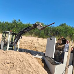 MaxMel - Koparka Terex opuszcza dużą betonową płytę do wykopu wyłożonego czarną folią, obok pracownika i usypanej ziemi, pod bezchmurnym niebem.