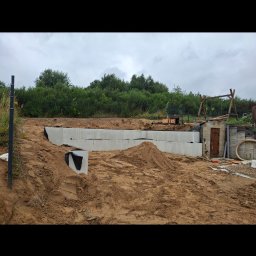 MaxMel - Ułożone bloczki betonowe na wykopie pod fundamenty, otoczone piaszczystym gruntem z widocznymi hałdami ziemi i zielonymi krzewami w tle, pod zachmurzonym niebem.