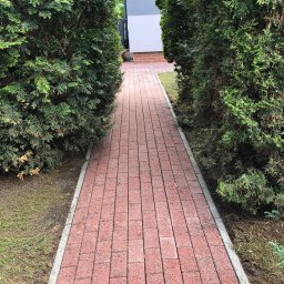 Filip Gawroński - Czerwona kostka brukowa tworzy ścieżkę w ogrodzie, otoczoną zielonymi tujami. Widok z perspektywy pieszego, prowadzący do domu z elementami cegły.