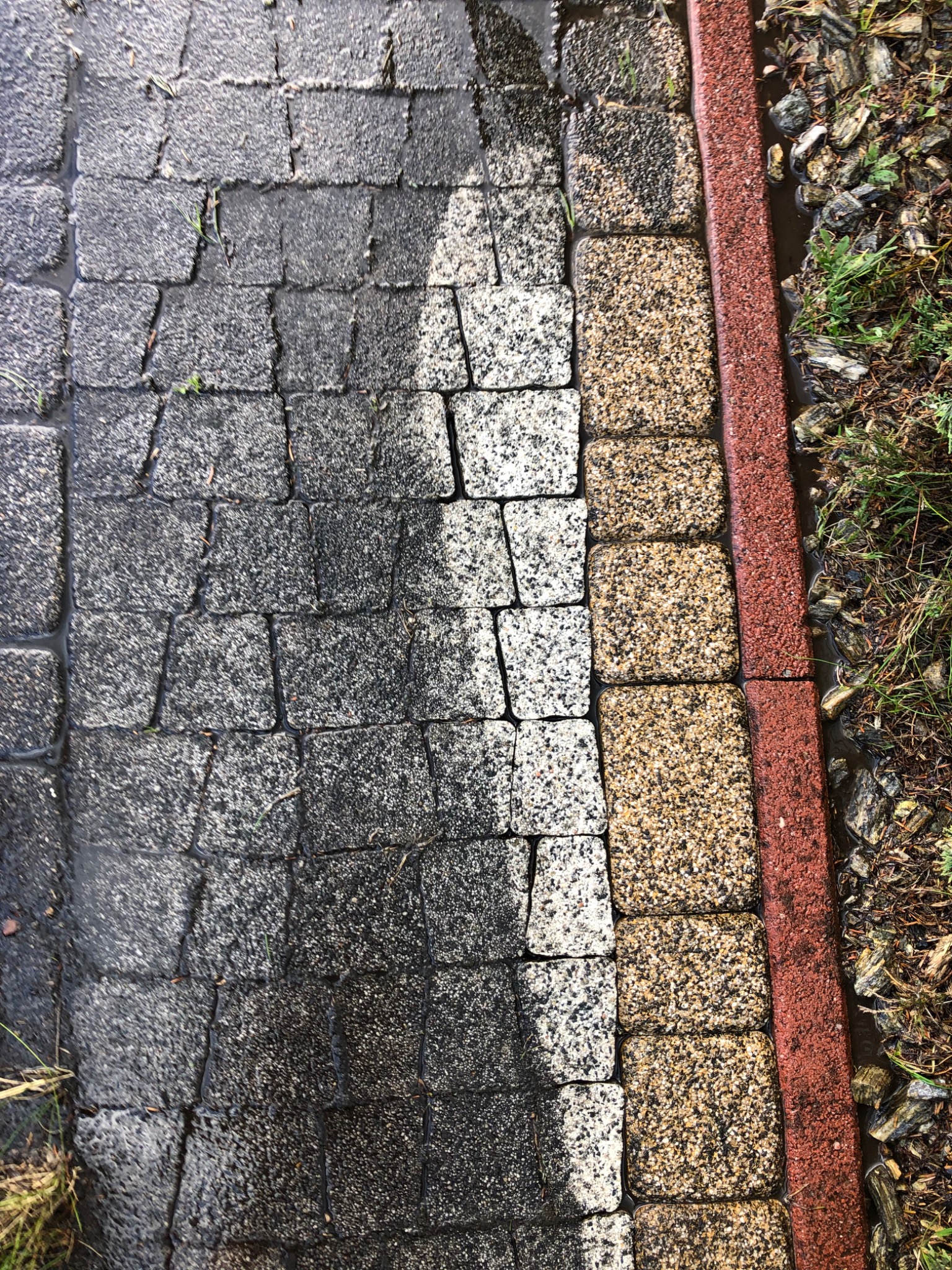 Częściowo umyta, mokra kostka brukowa w odcieniach szarości i bieli, obok kostka w kolorze piaskowym, oddzielona czerwoną obwódką, a dalej ziemia z roślinnością.