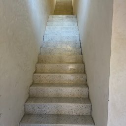 MIMAR Marek Perek - Klatka schodowa z terazzo, widok z dołu. Ściany w jasnym odcieniu beżu, schody w odcieniach szarości. Ujęcie centralne, symetryczne, z widocznym oświetleniem na górze.