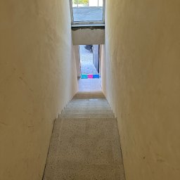 MIMAR Marek Perek - Klatka schodowa w trakcie prac wykończeniowych: ściany po szpachlowaniu, schody wyłożone płytkami z terazzo, widok z góry na dół, w tle okno i wejście.