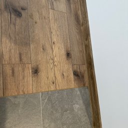 MIMAR Marek Perek - Połączenie podłogi: panele drewnopodobne i szare płytki. Widoczna biała ściana i drewniana listwa przypodłogowa. Ujęcie z góry, ukazujące detale materiałów.