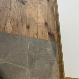 MIMAR Marek Perek - Połączenie podłogi z paneli drewnopodobnych i szarych płytek, wykończone listwą przypodłogową. Widoczny fragment węża odkurzacza.