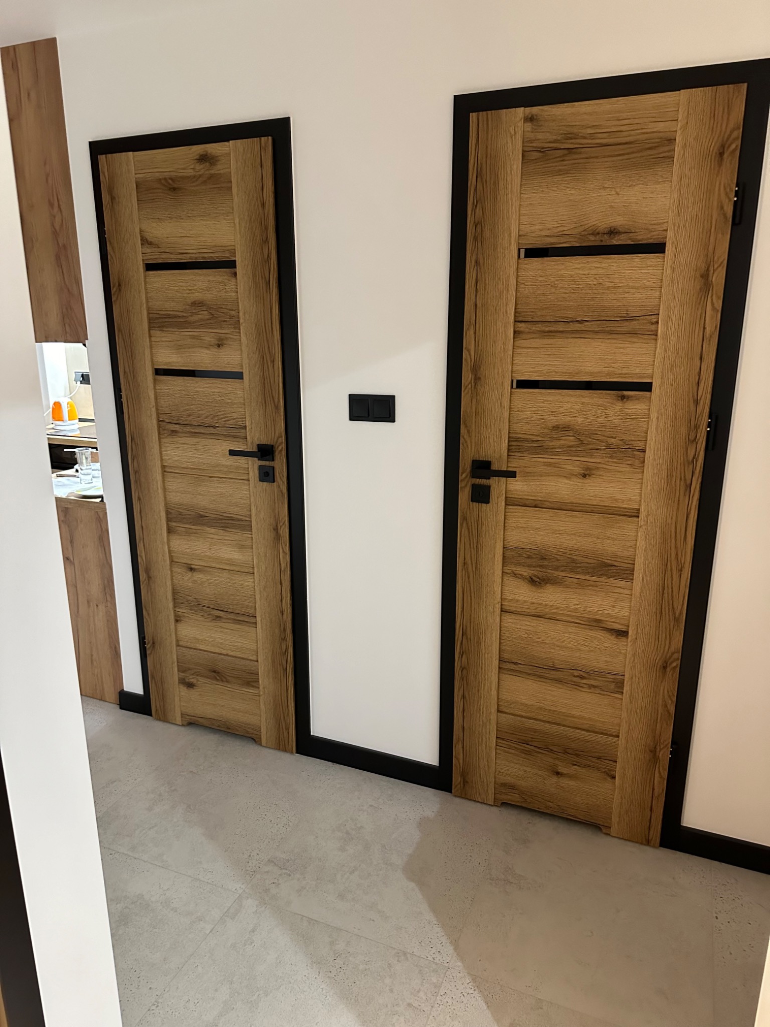 Dwa drewniane drzwi z czarnymi ramami i klamkami, widoczne na tle białej ściany i szarej podłogi. W tle widoczny fragment pomieszczenia z wyposażeniem.