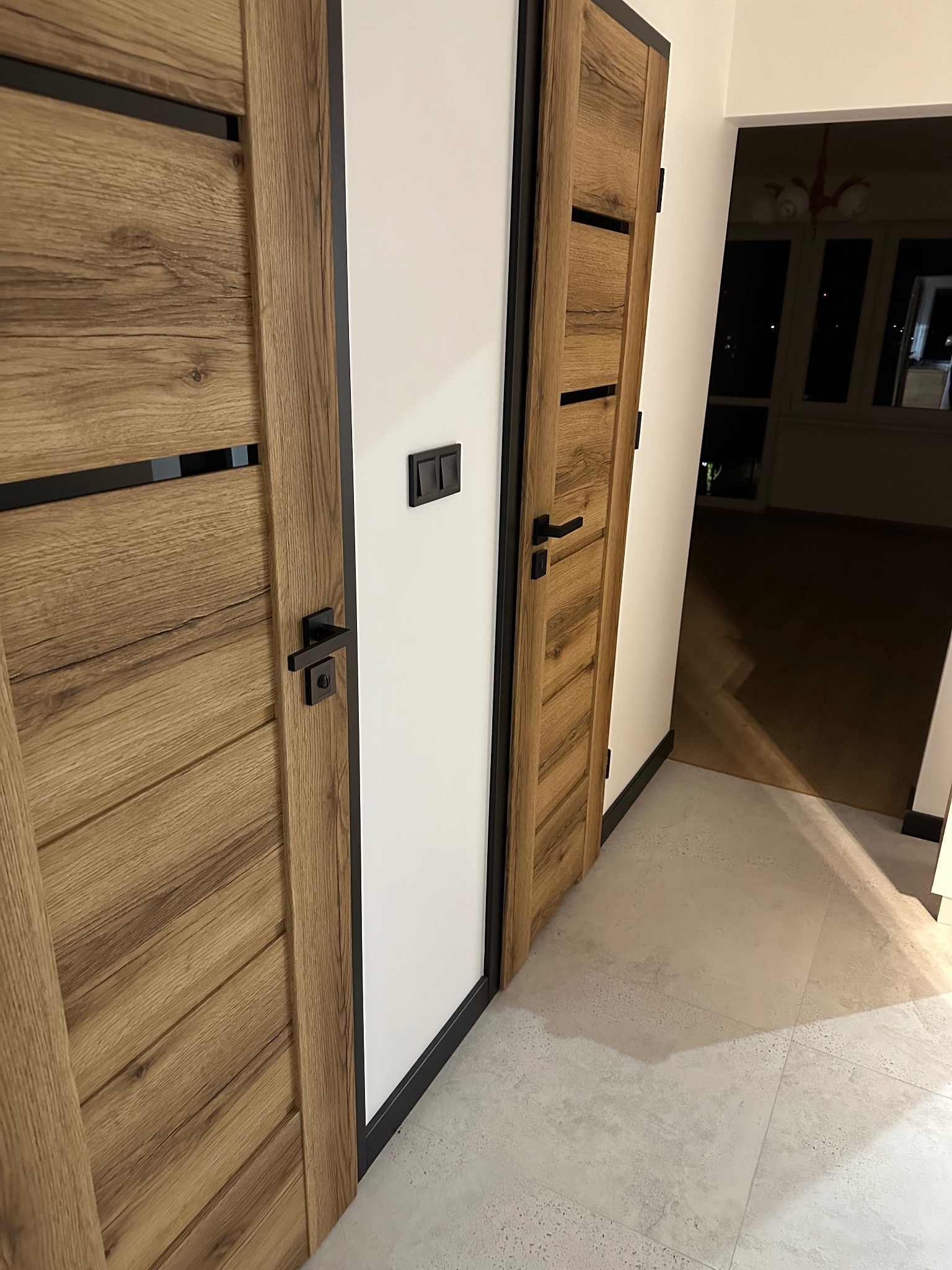Dwa drewniane drzwi z czarnymi detalami, widoczne w korytarzu z szarą podłogą. W tle widoczny balkon z oknami i lampą.