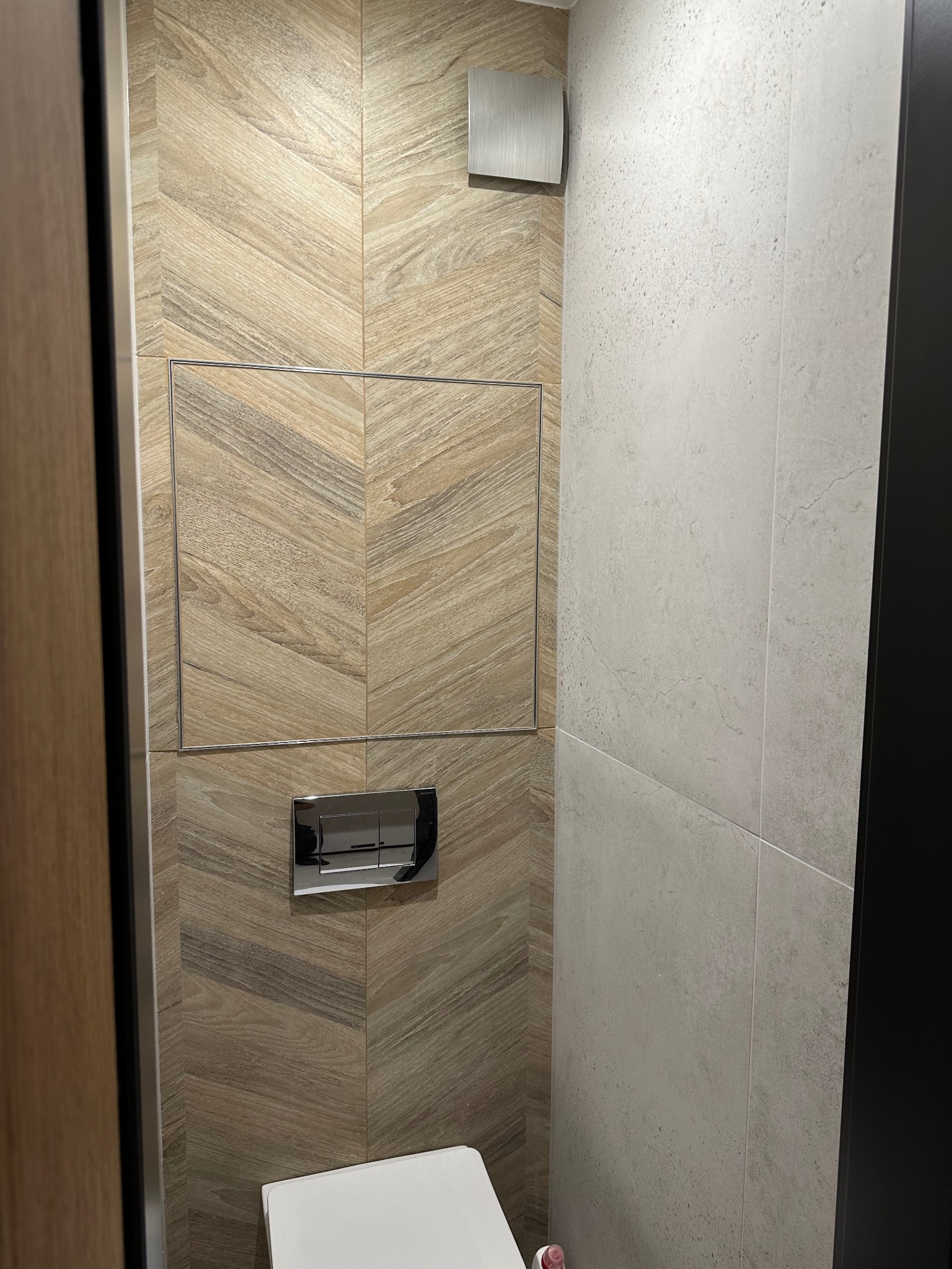 Wykończona łazienka z toaletą podwieszaną i płytkami imitującymi drewno ułożonymi w jodełkę. Widoczny rewizyjny właz i minimalistyczny przycisk spłukujący.