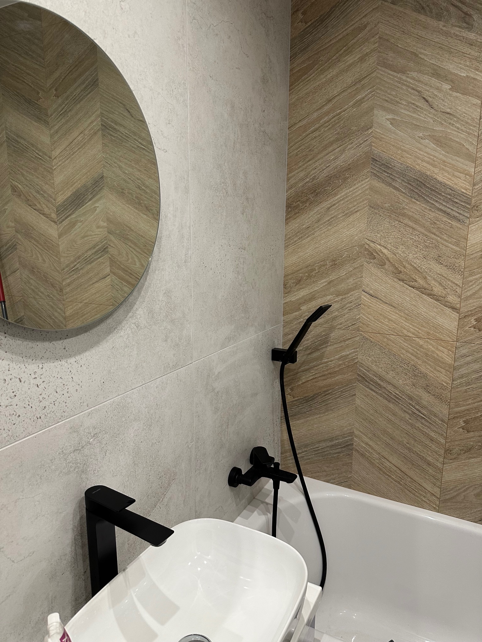 Nowoczesna łazienka z okrągłym lustrem, białą umywalką i czarną armaturą na tle płytek imitujących drewno ułożonych w jodełkę.