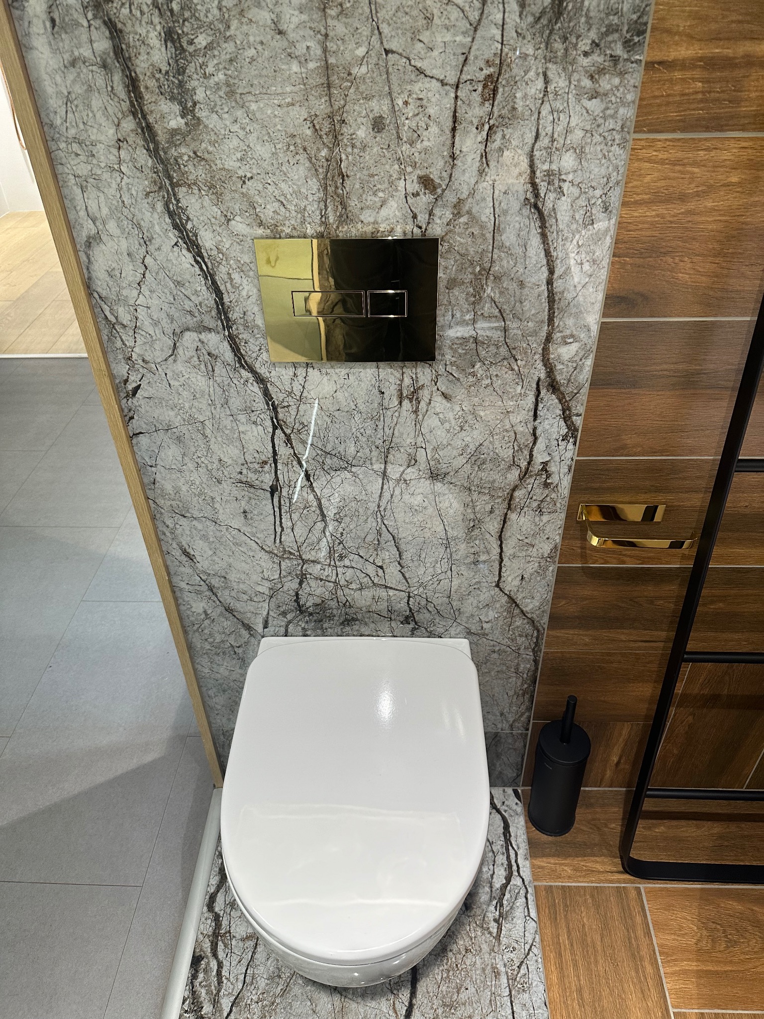 Nowoczesna toaleta w wykończonej łazience. Marmurowe płytki na ścianie z złotym przyciskiem spłuczki, drewnopodobne płytki na ścianie obok. Widoczny fragment drabinki.