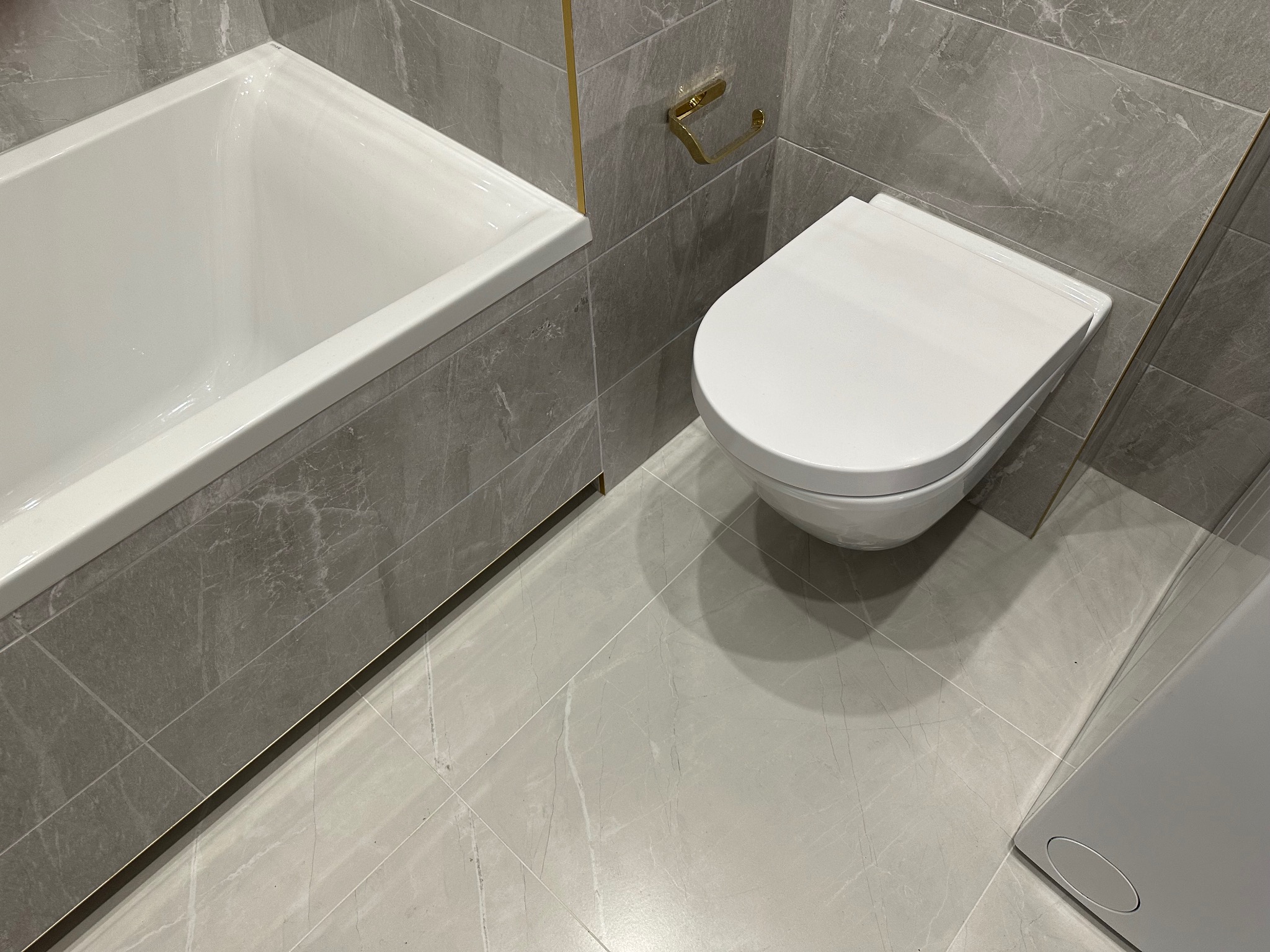 Nowoczesna łazienka w szarościach: marmurowe płytki, biała wanna i podwieszana toaleta z akcentami złota. Minimalistyczny design, eleganckie wykończenie.