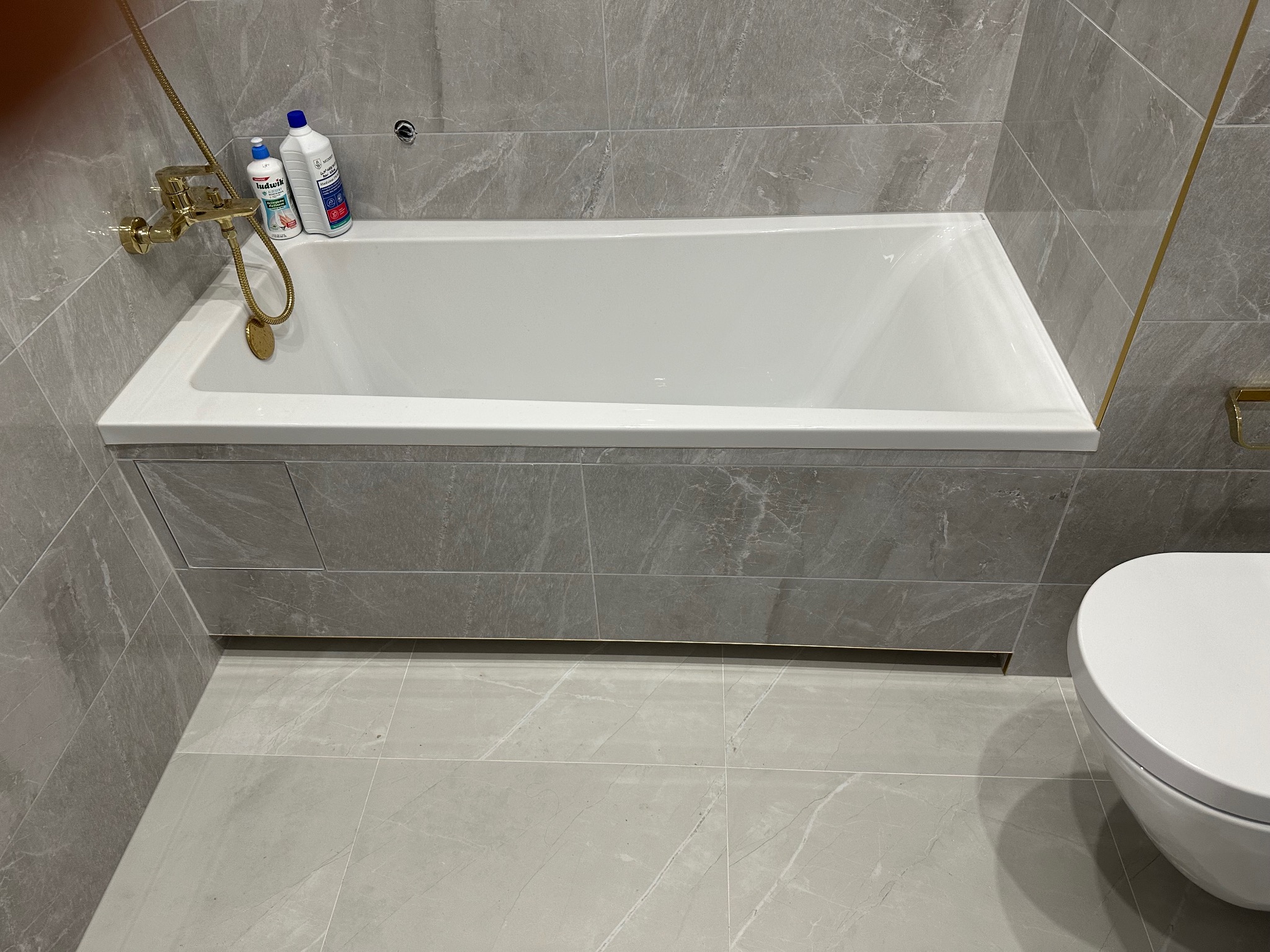 Nowoczesna łazienka z wanną obudowaną płytkami imitującymi marmur, złota armatura i akcesoria, szare płytki na ścianach i podłodze, toaleta w tle.